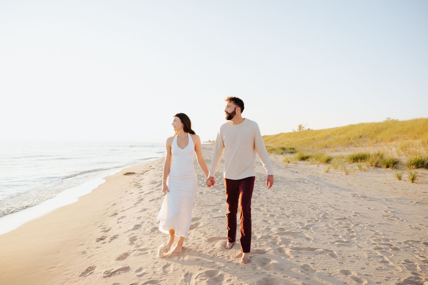 A couple walks hand-in-hand along a sunny sandy beach, wearing white outfits, with ocean waves and green dunes behind them.