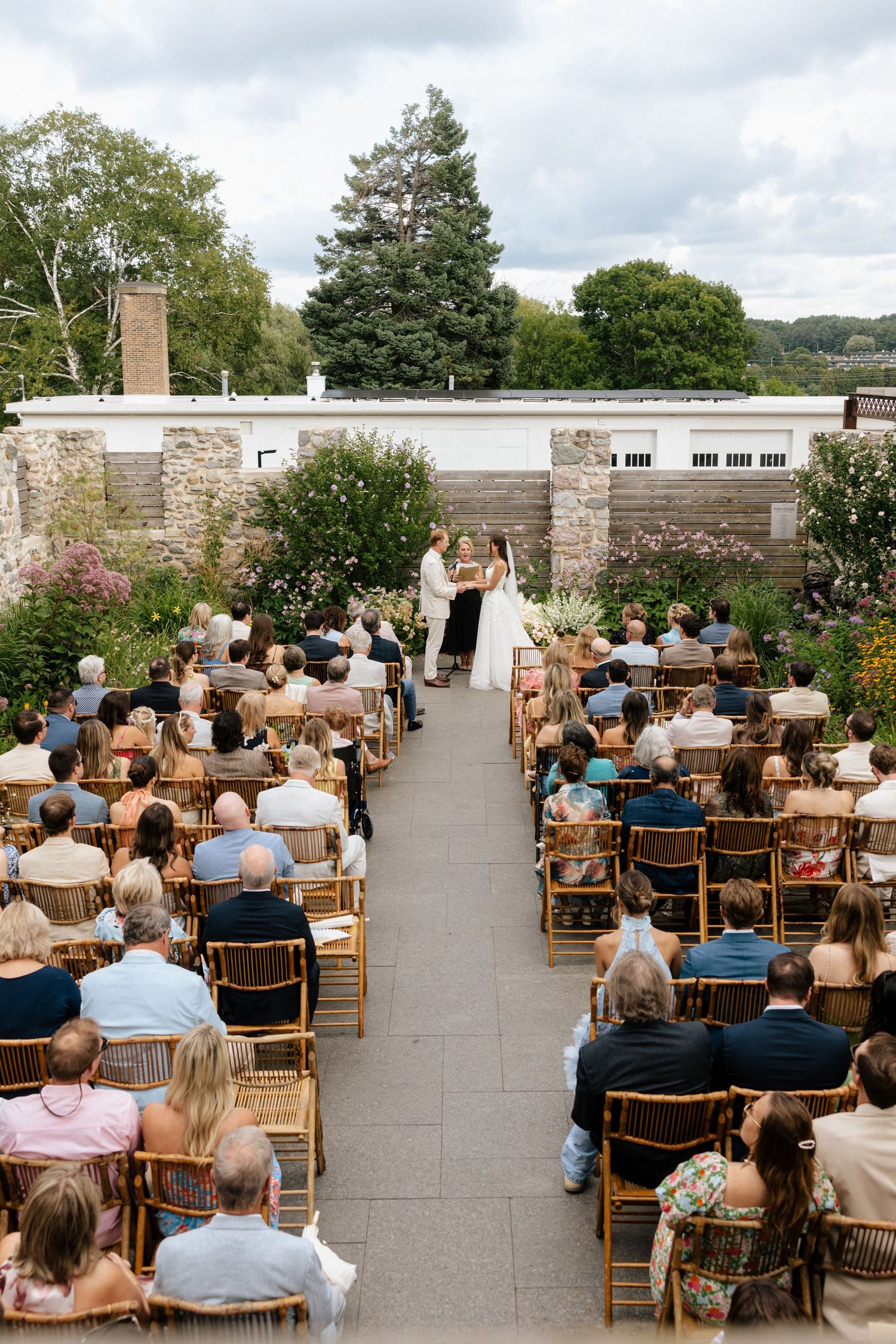 Outdoor wedding ceremony with guests seated in wooden chairs along an aisle leading to couple at altar amid lush gardens.