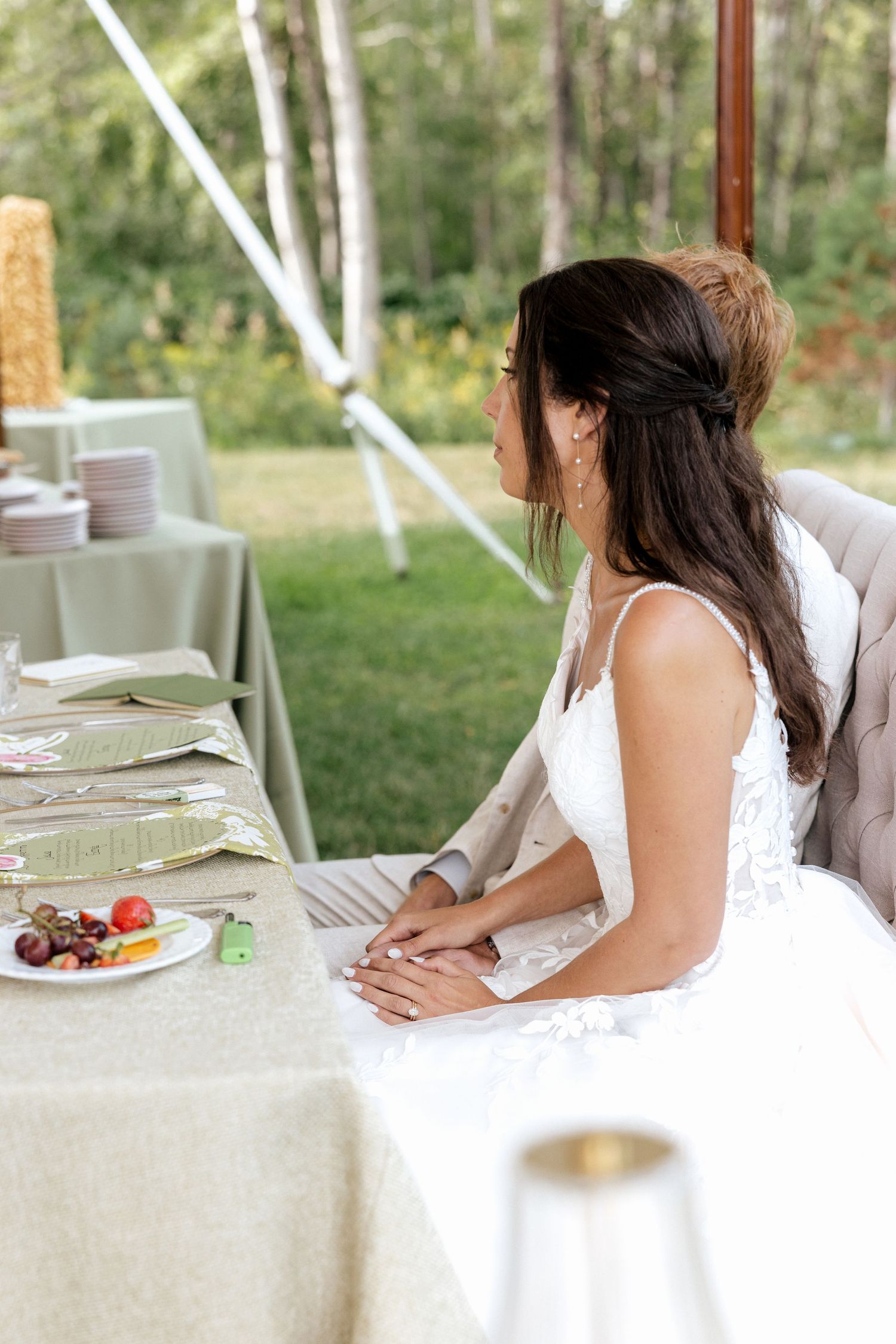 Bride in lace wedding gown seated at reception table with menu cards and fruit plate.