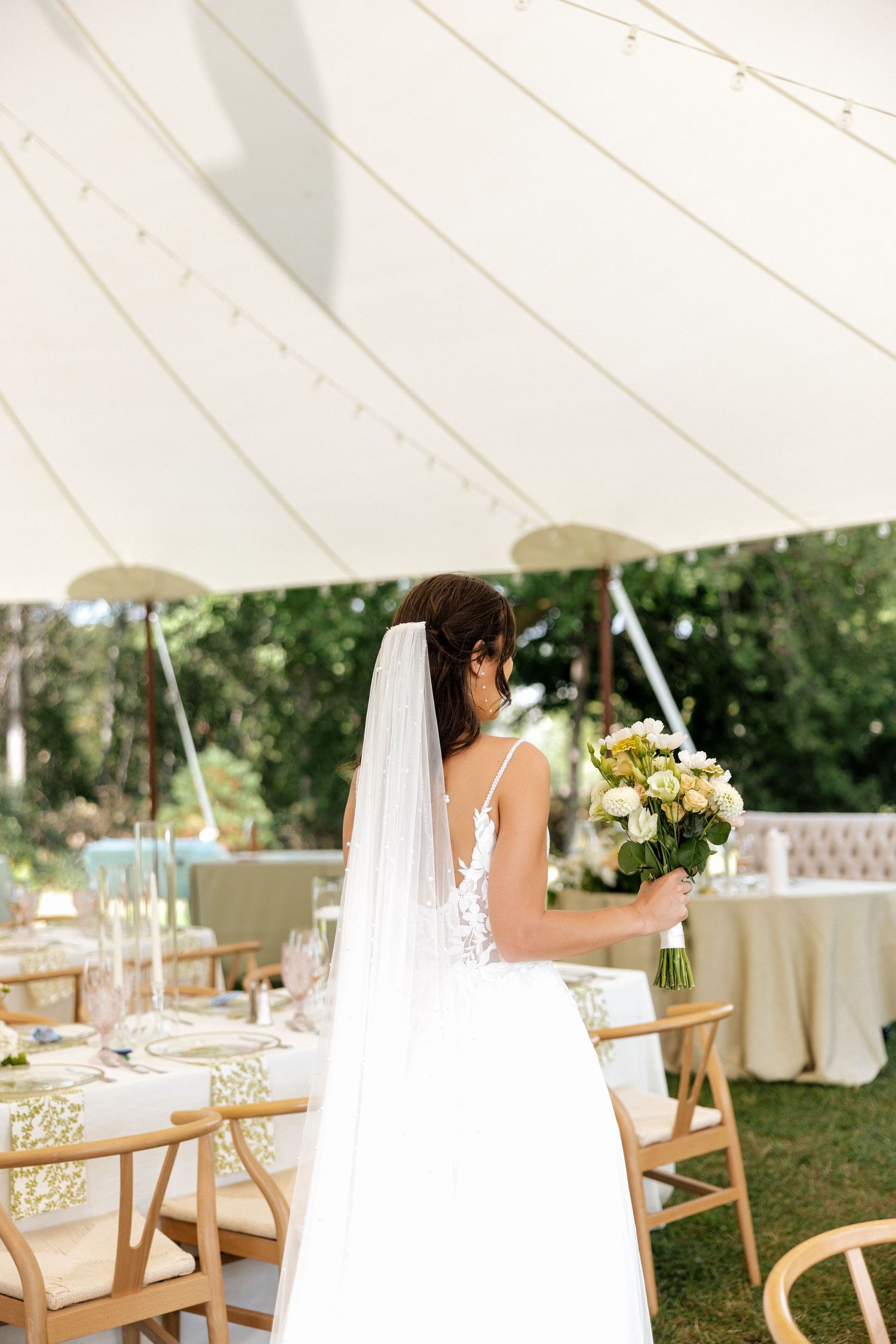 Bride in white lace gown and veil holding bouquet inside elegant outdoor wedding tent.