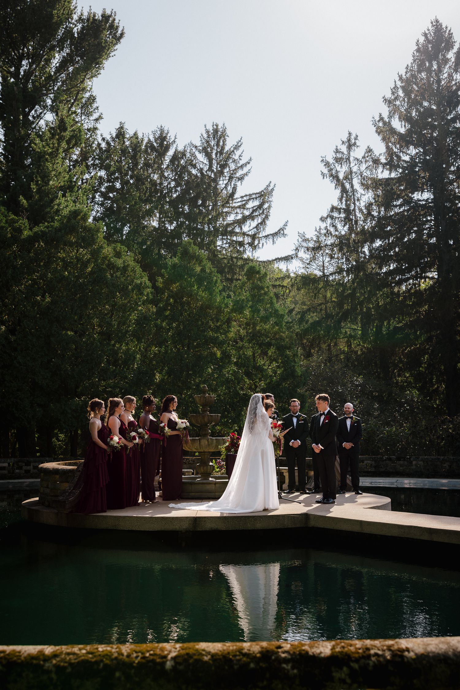 Outdoor wedding ceremony by a reflective pool, bride in white gown with bridesmaids in burgundy dresses, surrounded by tall trees.
