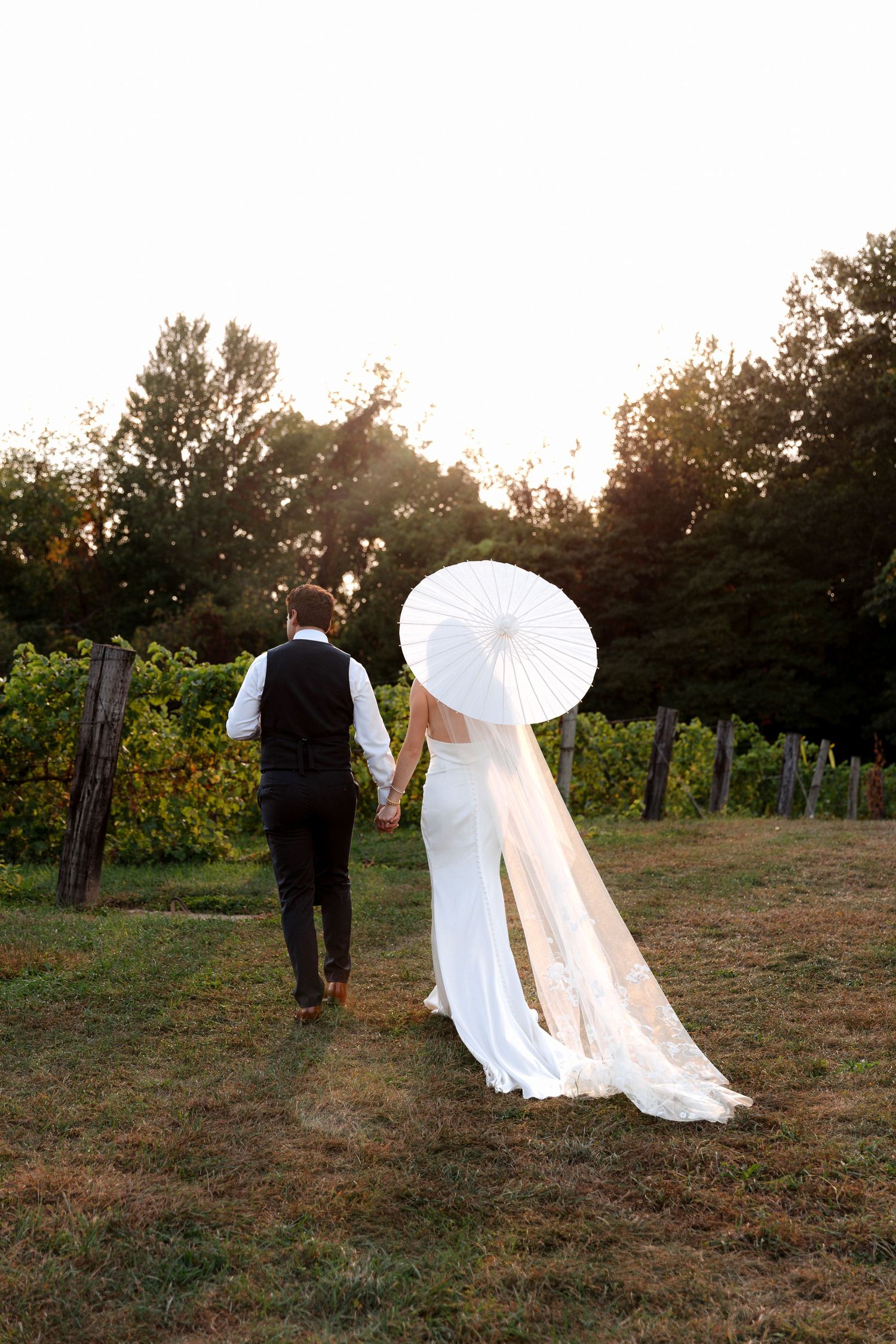 Bride and groom walking hand-in-hand through a vineyard at sunset, bride holding a white parasol with long veil trailing.