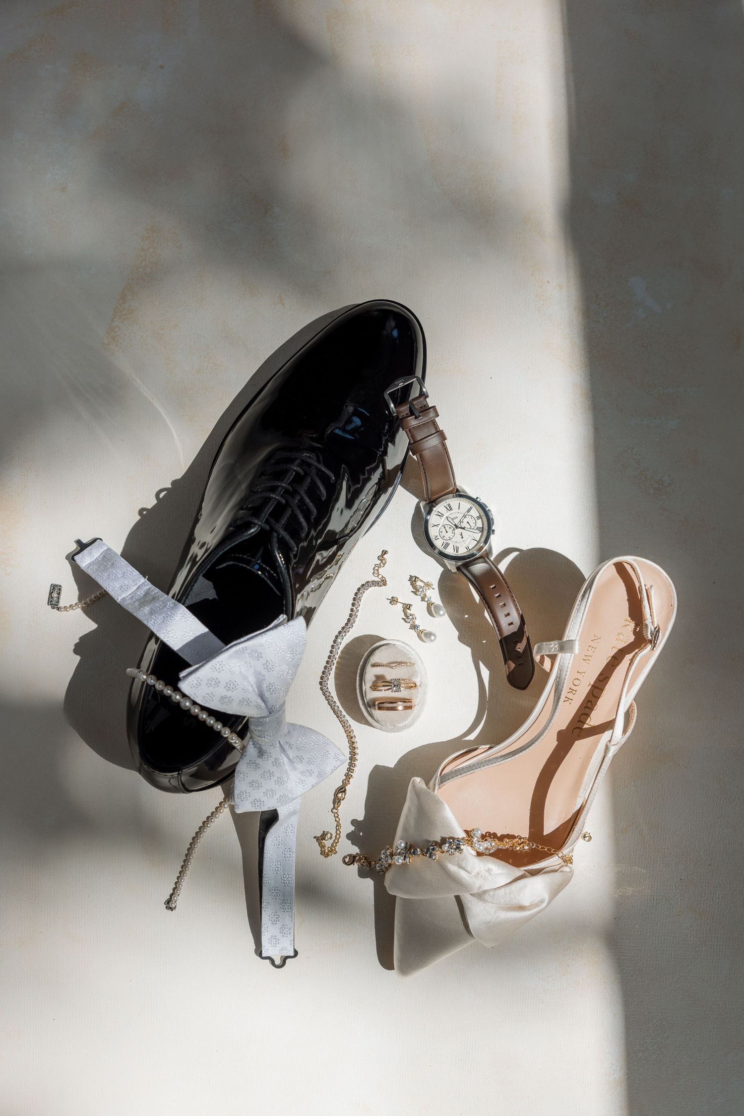 Wedding accessories flat lay featuring black dress shoes, strappy heels, a watch, garter, rings, and jewelry on a marble surface.