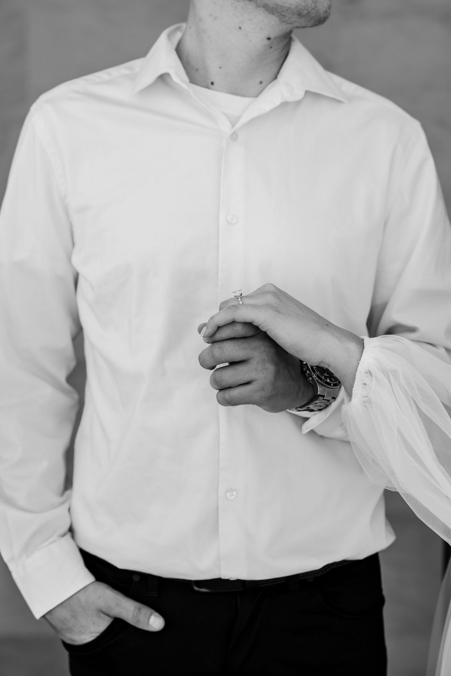 Black and white photo of a groom in a white dress shirt holding hands with bride, wearing a watch.
