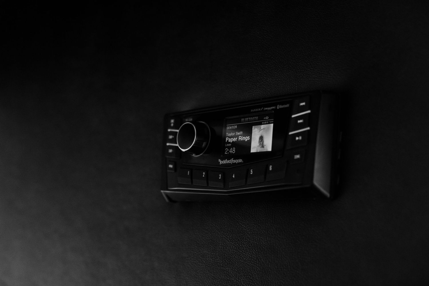 Black Sirius XM satellite radio receiver mounted on a dark wall with illuminated display screen.