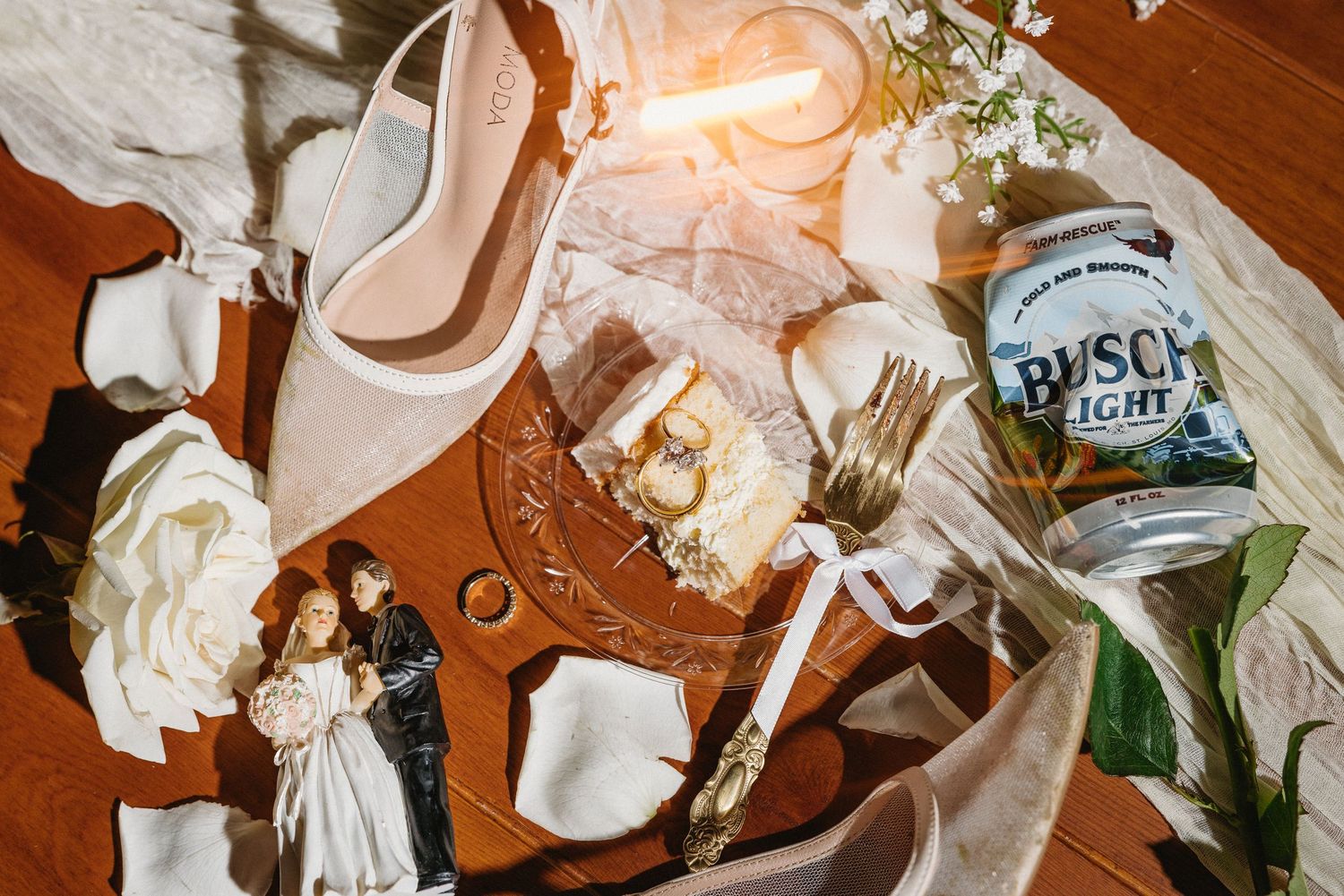 Wedding flat lay featuring bridal heels, cake topper, rings, white roses, and a Busch Light beer can.
