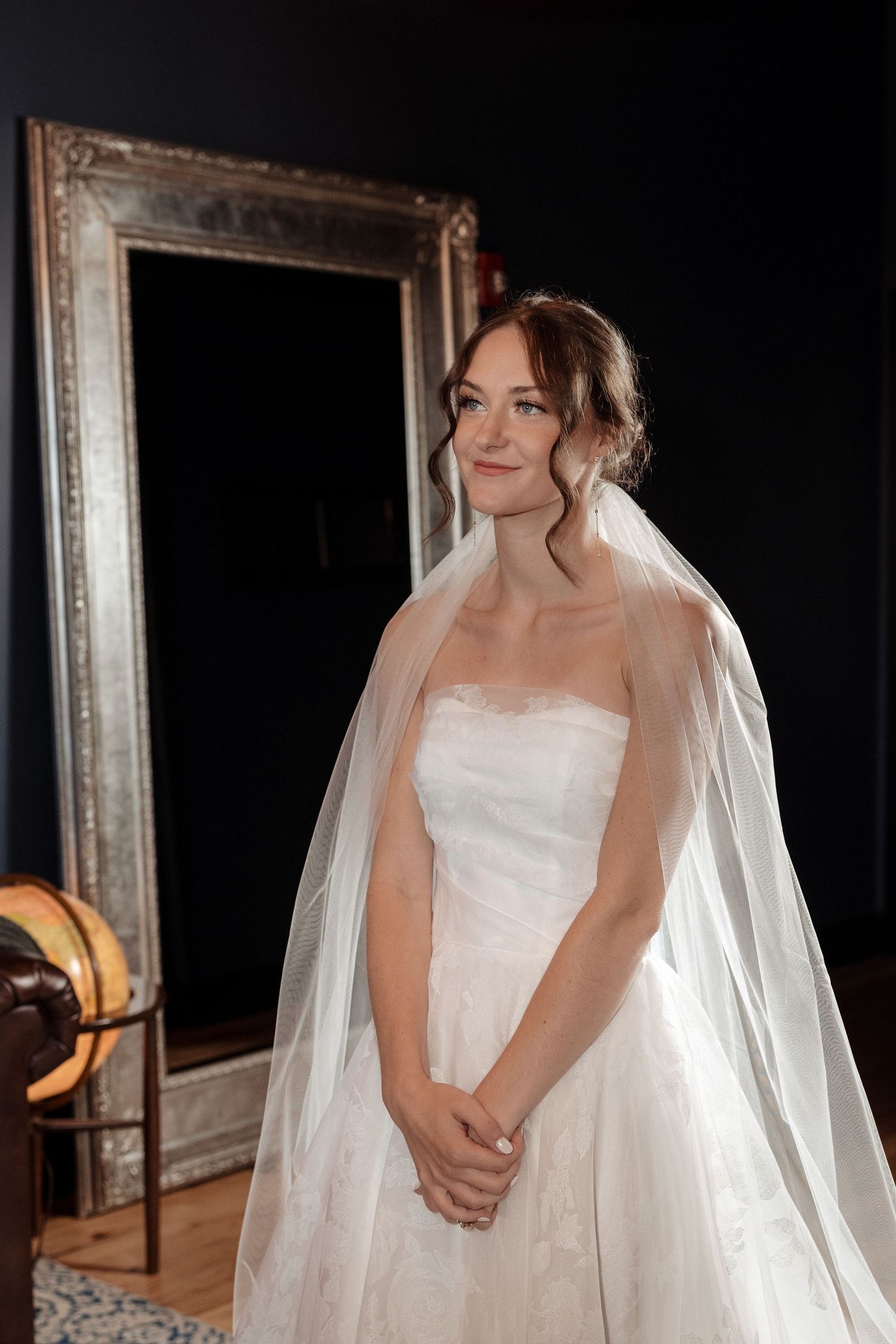 Elegant bride in a strapless white lace gown and veil poses before a large ornate mirror indoors.