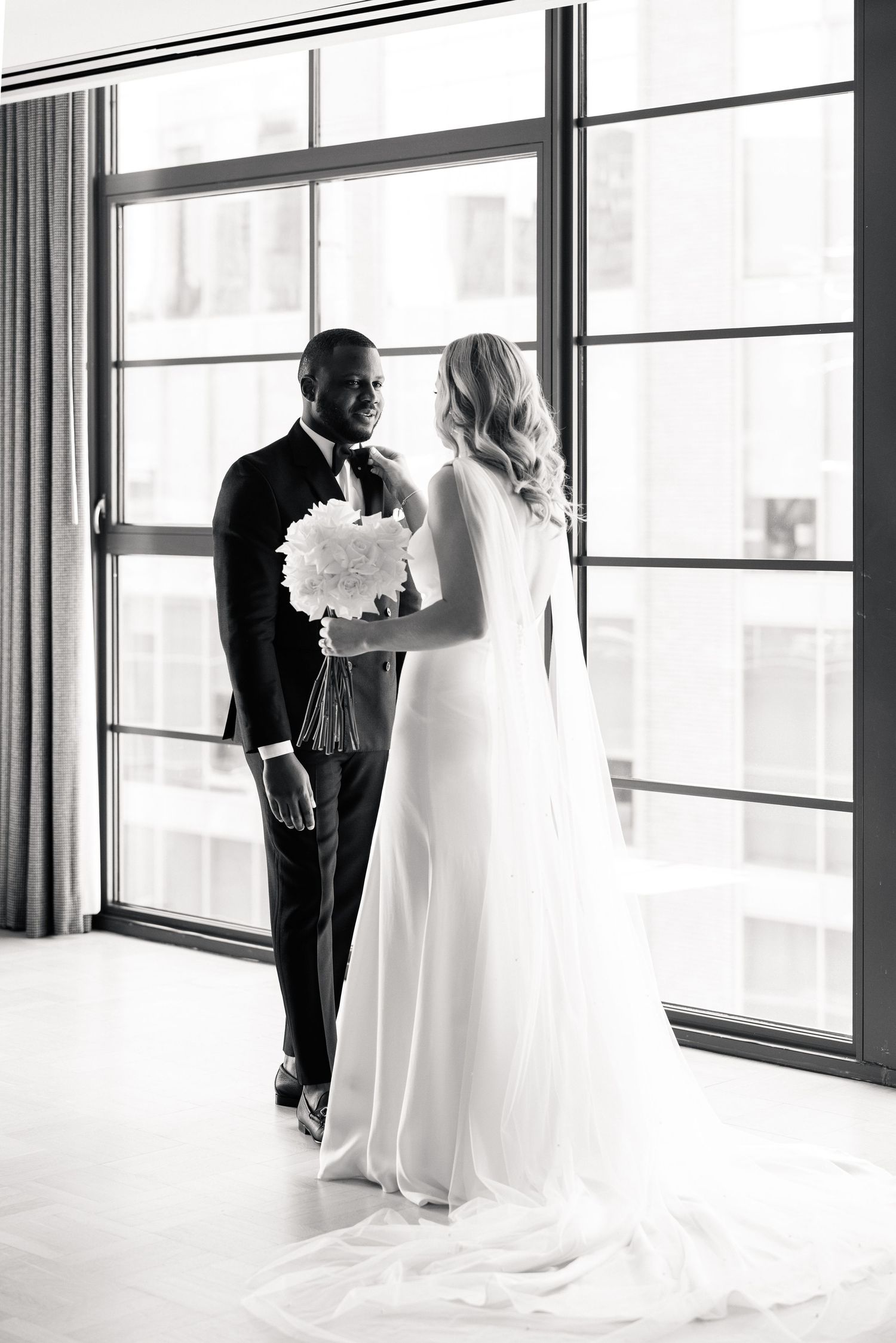 Black and white wedding portrait of interracial couple sharing first look near floor-to-ceiling windows, bride holding bouquet.