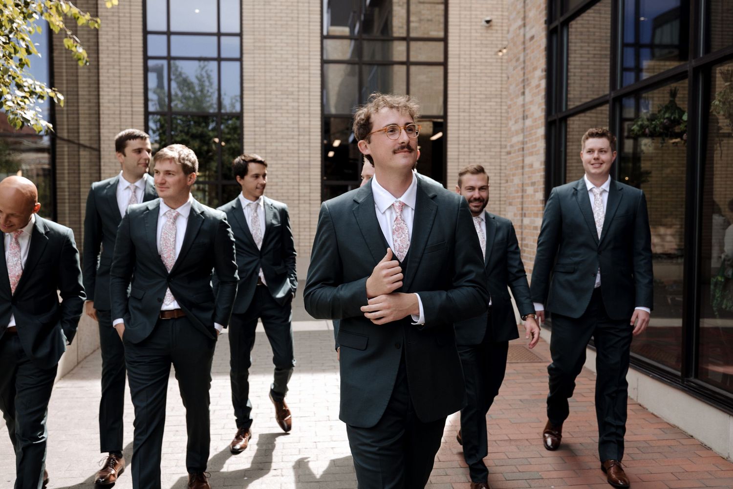Groomsmen in dark suits with pink ties walk confidently together outside a brick building on a wedding day.