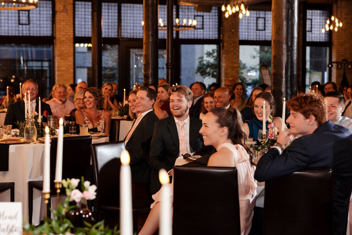 Wedding reception guests laughing and enjoying speeches at elegantly decorated tables in a modern brick venue.