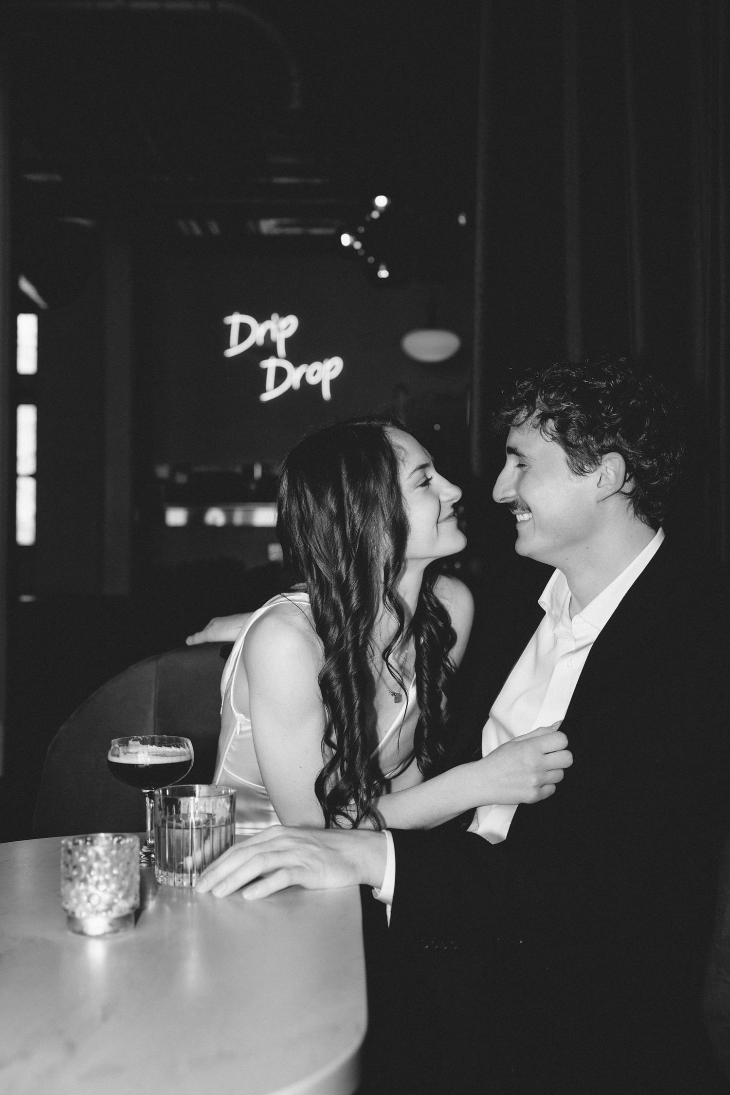 Black and white photo of a romantic couple smiling at each other at a bar with a Drip Drop neon sign.