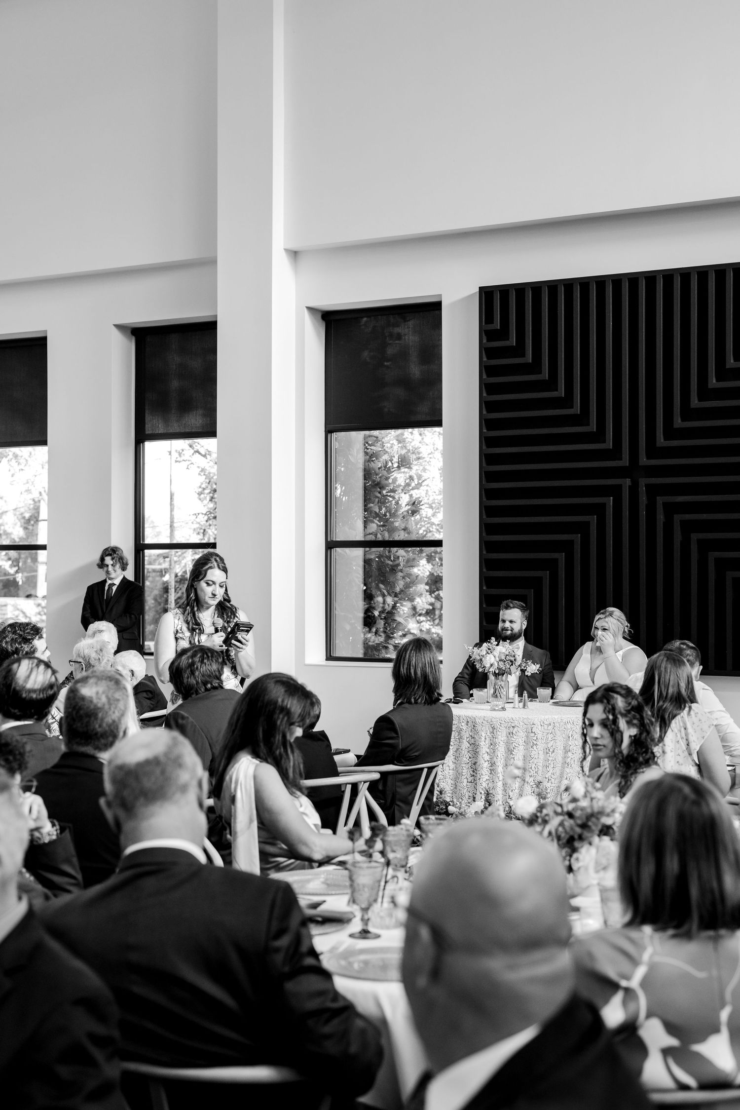 Black and white photo of a woman giving a speech at an elegant wedding reception dinner with guests seated at decorated tables.