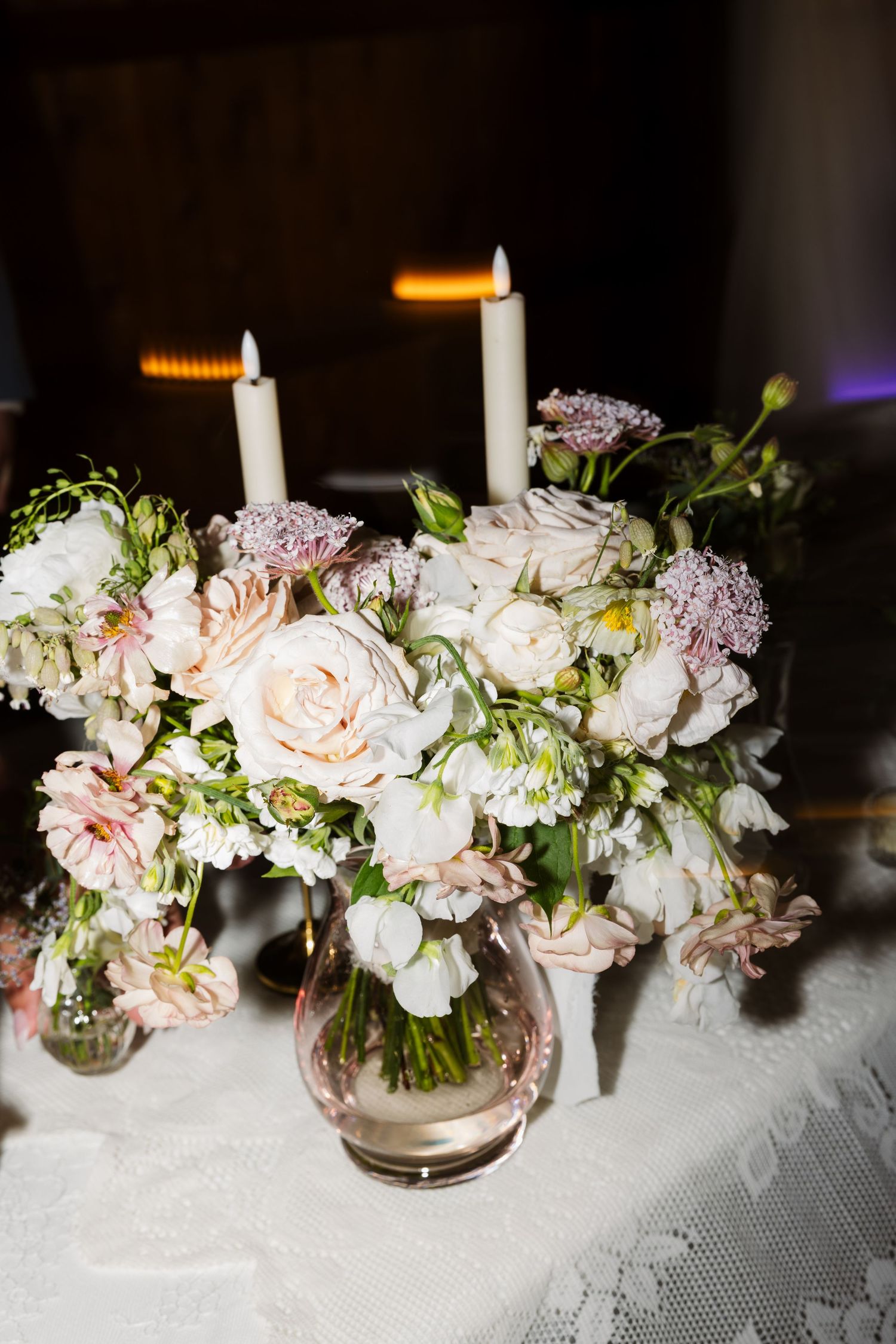 Elegant wedding centerpiece with white and blush roses in glass vase, accented by lit taper candles on lace tablecloth.