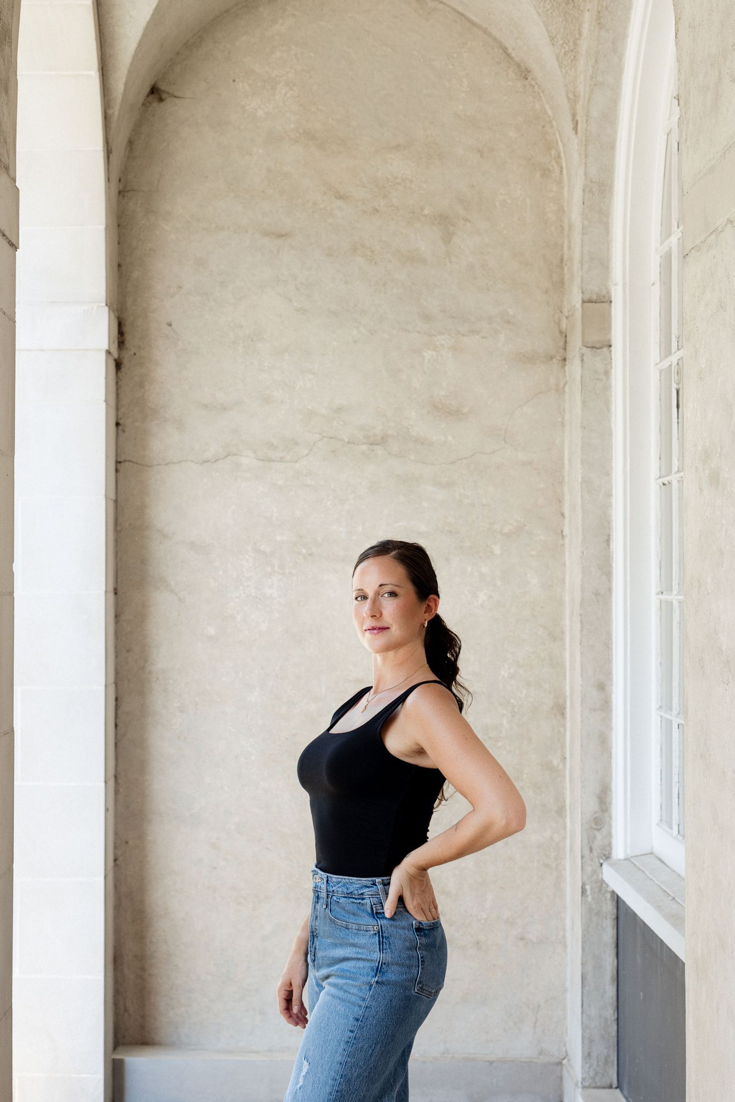 Woman in black tank top and blue jeans posing confidently under stone arches in a classic architectural setting.