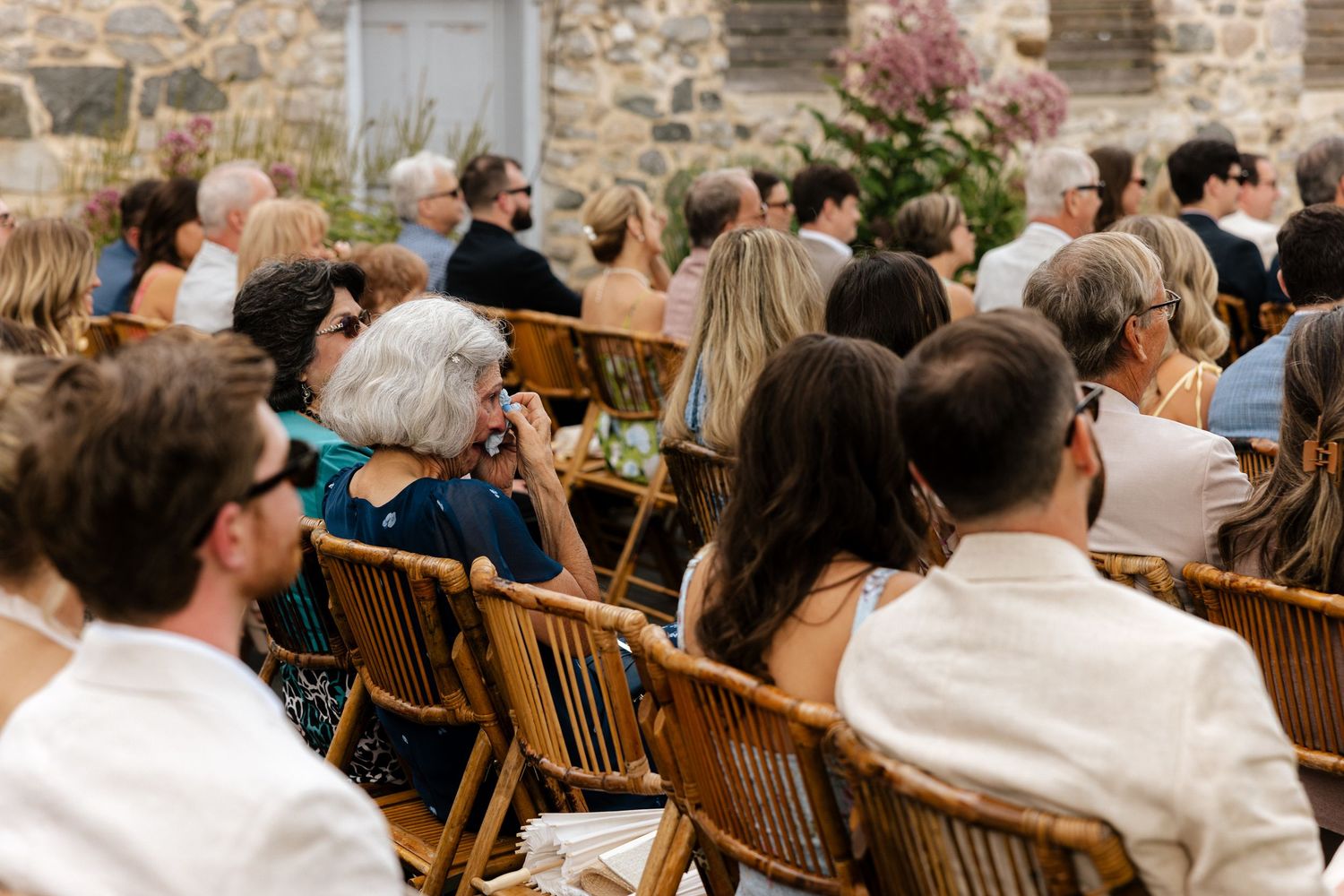 Guests seated in wooden chairs at an outdoor wedding ceremony, dressed in elegant attire.