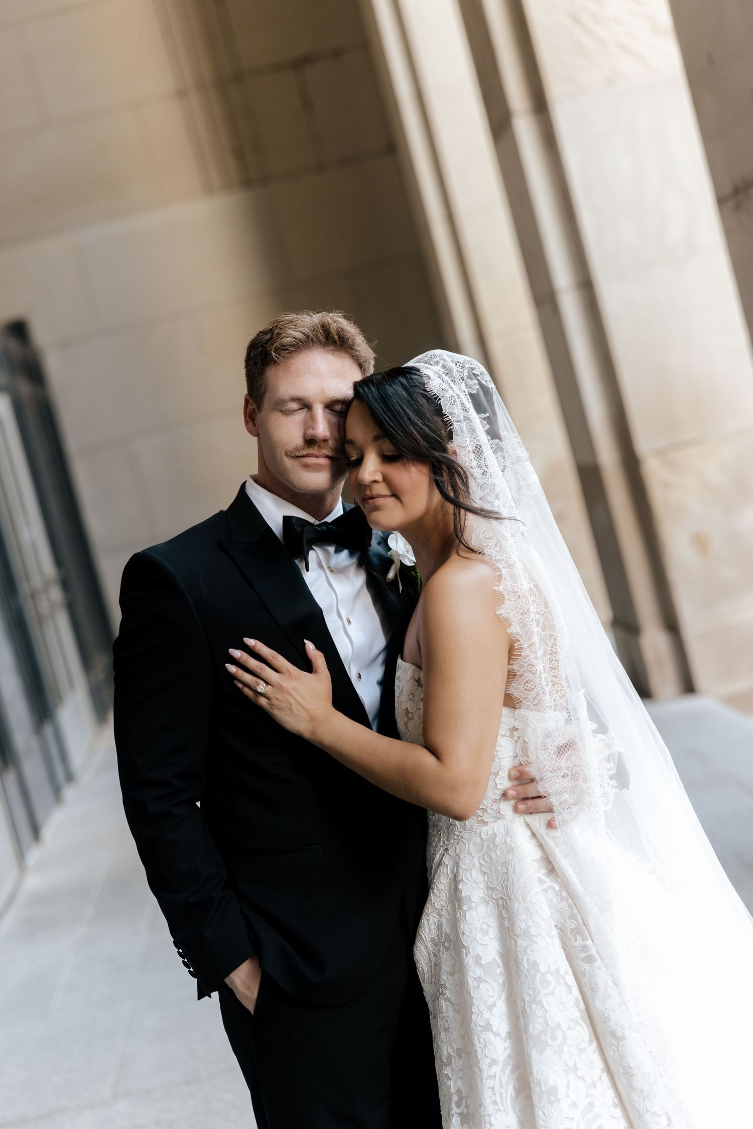 Elegant bride and groom embrace outdoors, wearing lace wedding gown with veil and classic black tuxedo with bow tie.