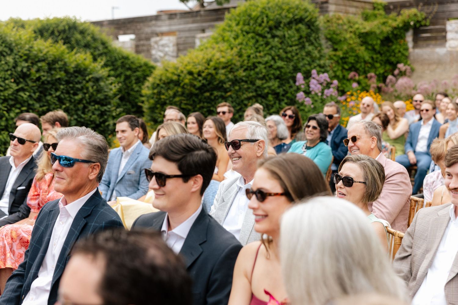 Guests wearing sunglasses seated outdoors at a sunny wedding ceremony, smiling and enjoying the celebration.
