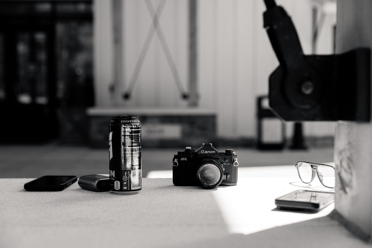 Black and white photo of a vintage camera, energy drink can, glasses, and phone on a table.