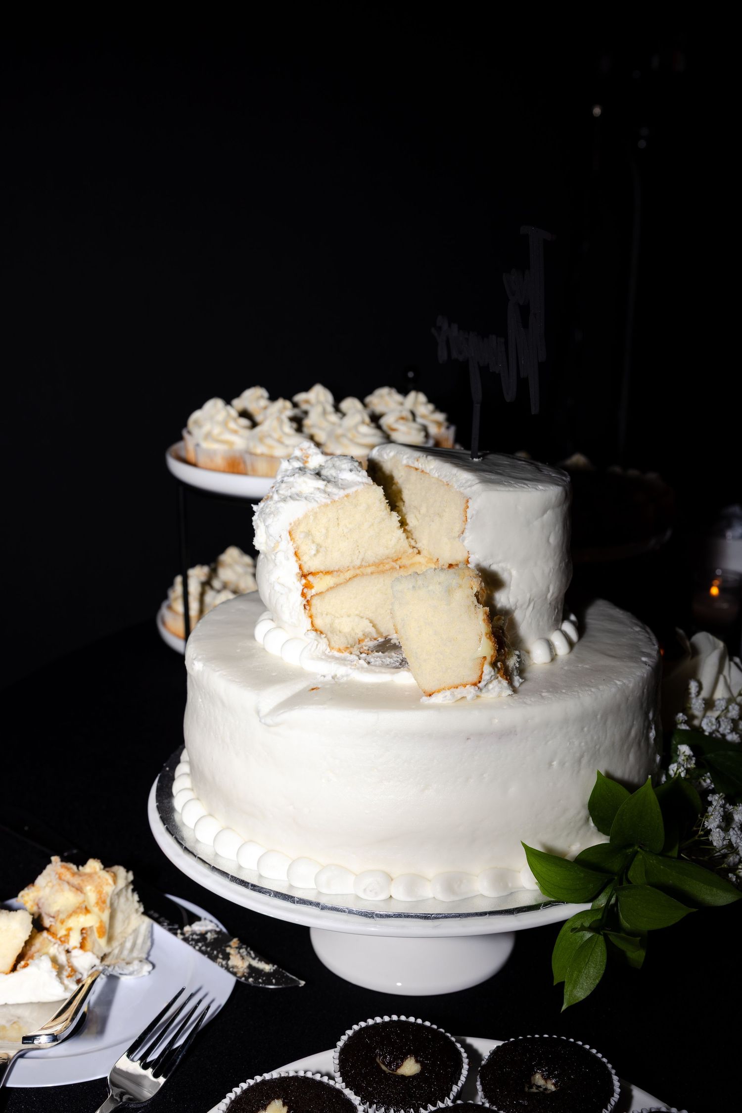 Two-tier white wedding cake with slices cut out, displayed on a cake stand with cupcakes and desserts in the background.