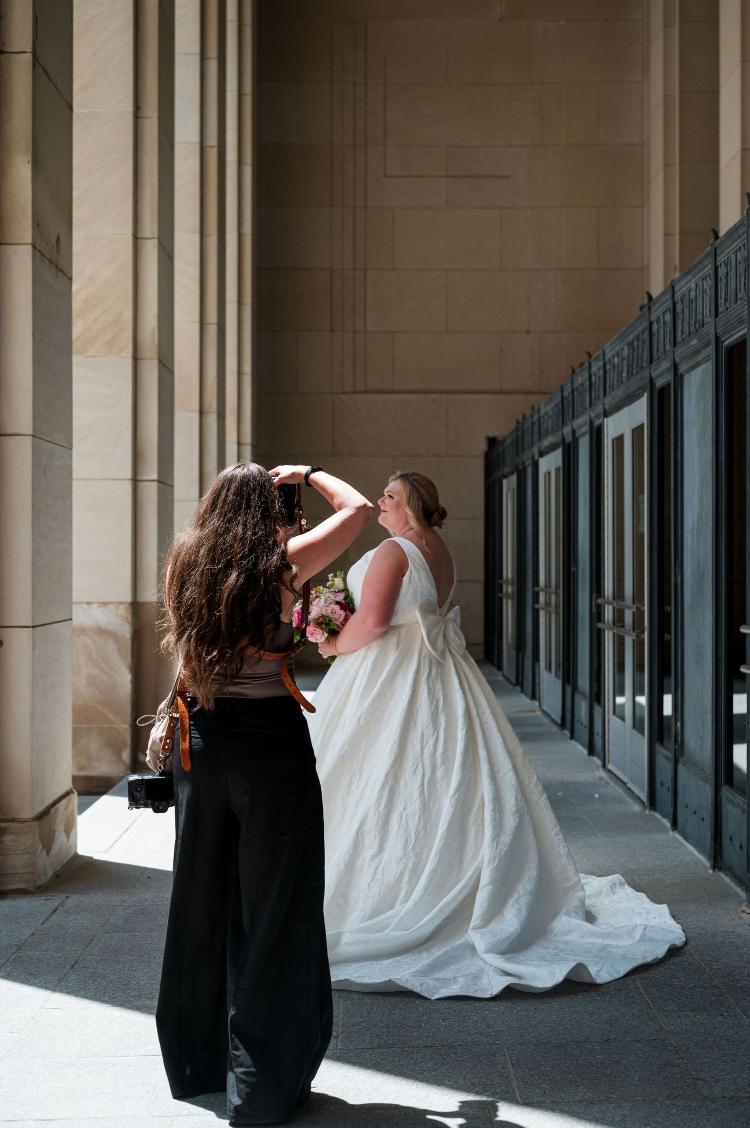Bride in white gown holds bouquet while assistant adjusts hair outside elegant stone building columns.