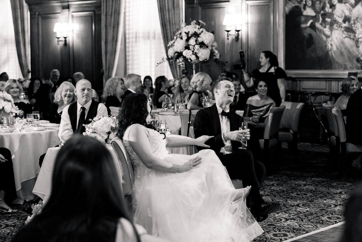 Black and white photo of bride and groom seated at wedding reception table surrounded by guests.