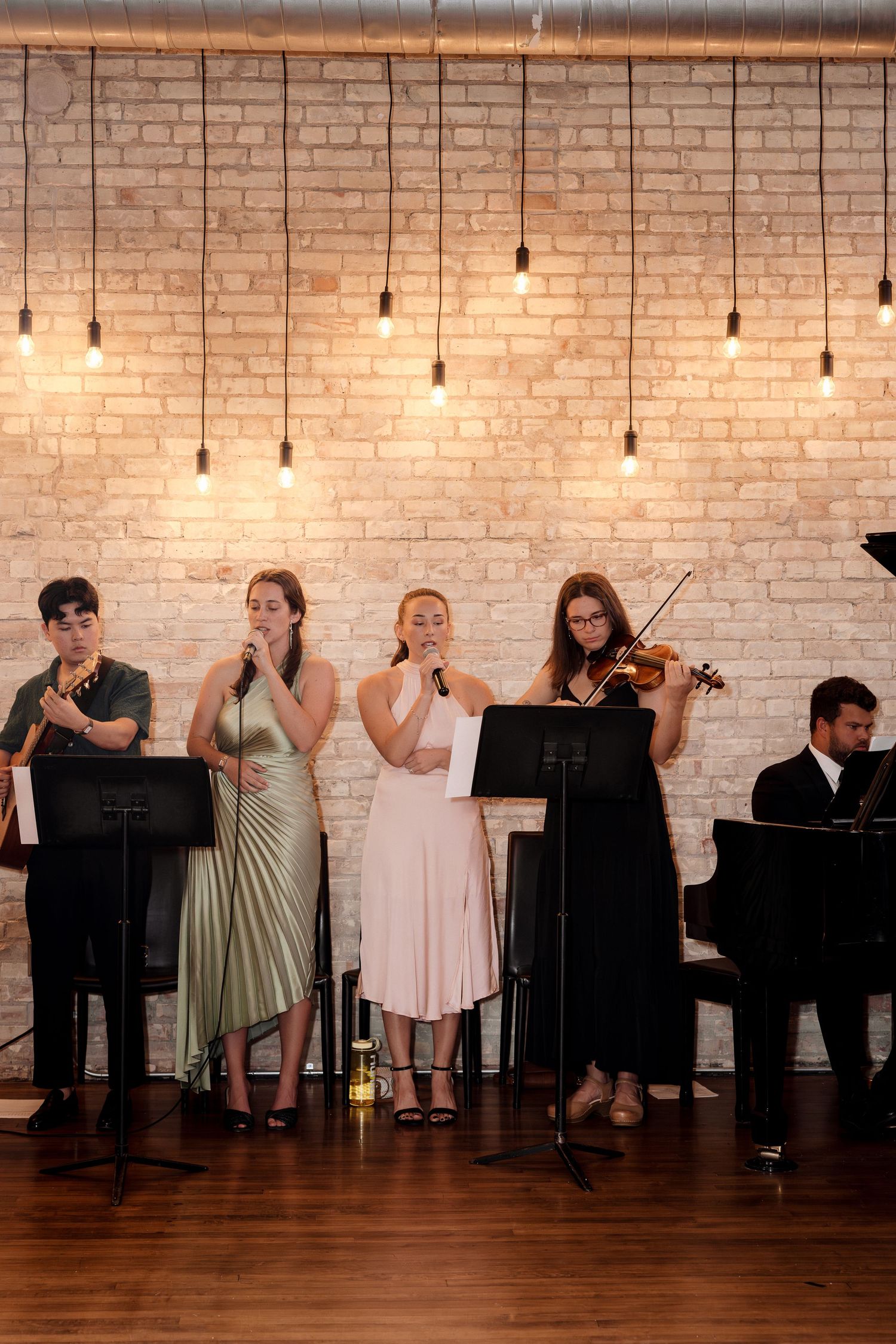 Live wedding band performs in front of a white brick wall with Edison bulb lighting at an elegant reception.