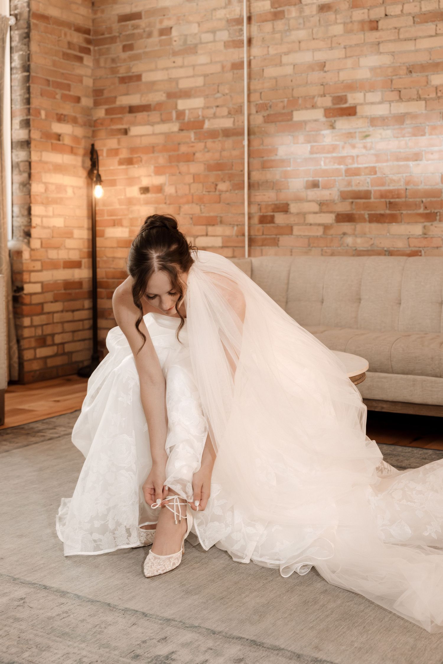Bride in a white gown with veil tying her lace heels in a brick-walled bridal suite.
