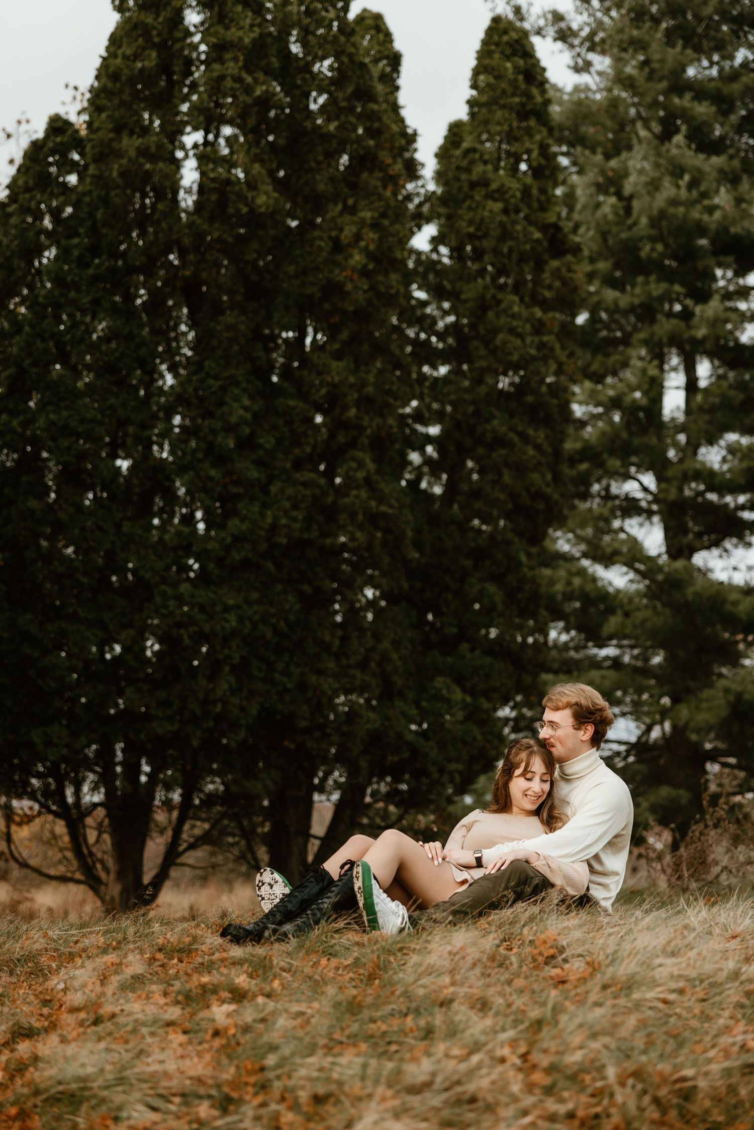 An engaged couple sits together laughing in a grassy field surrounded by tall evergreen trees.