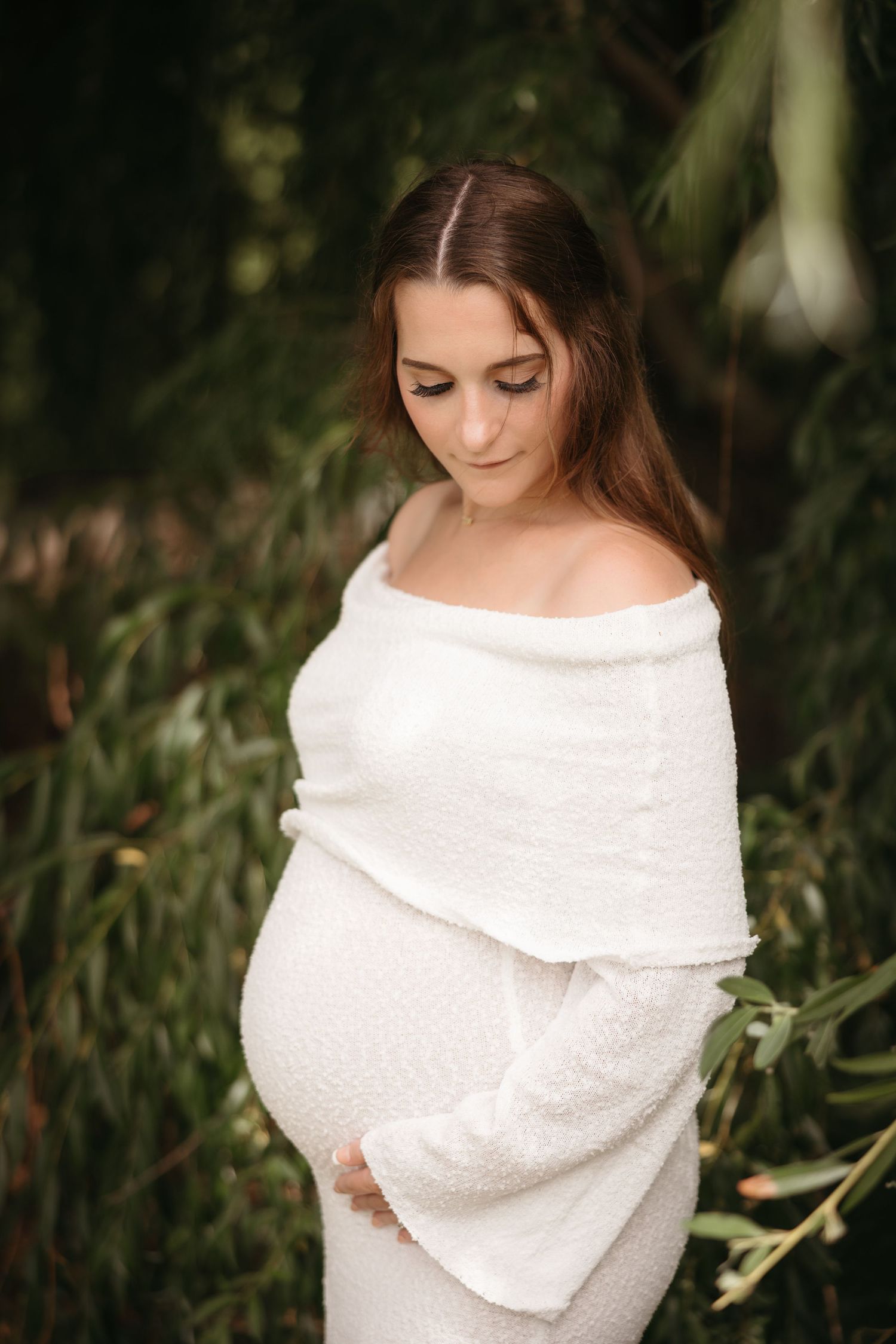 Serene pregnant woman in white off-shoulder dress posing among green foliage.