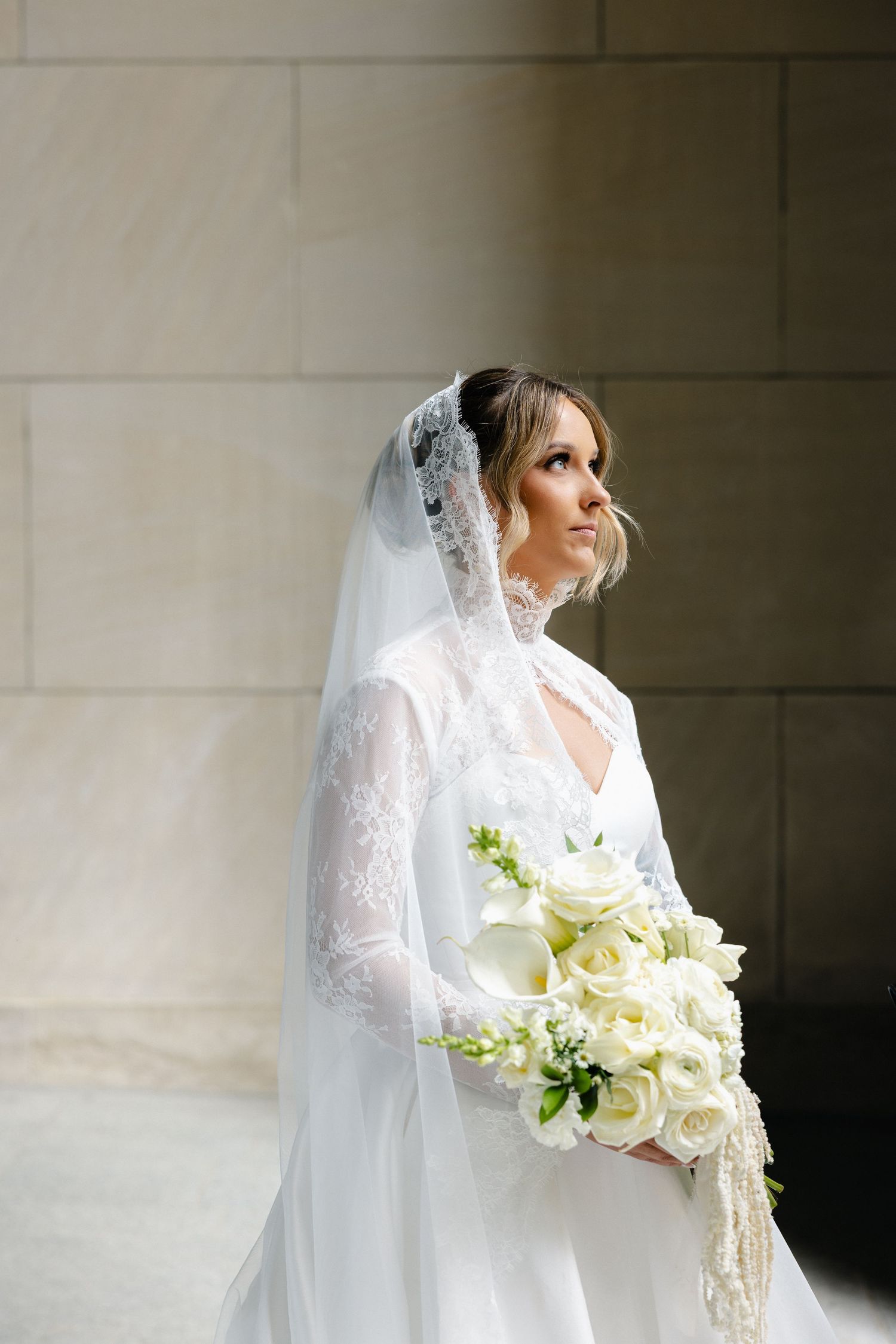 Elegant bride in lace veil and white gown holding bouquet of white roses and calla lilies.