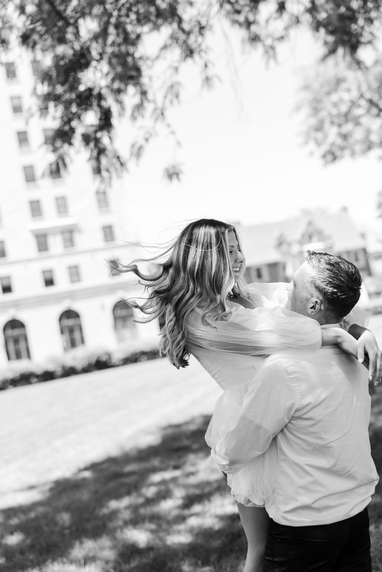 Black and white photo of a couple embracing outdoors, woman's hair flowing as man lifts her near a grand building.