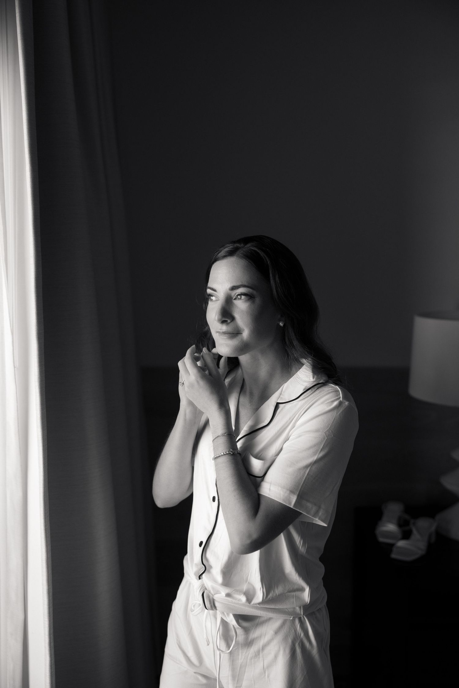 Black and white portrait of bride in pajamas standing by window in soft light during wedding morning prep.