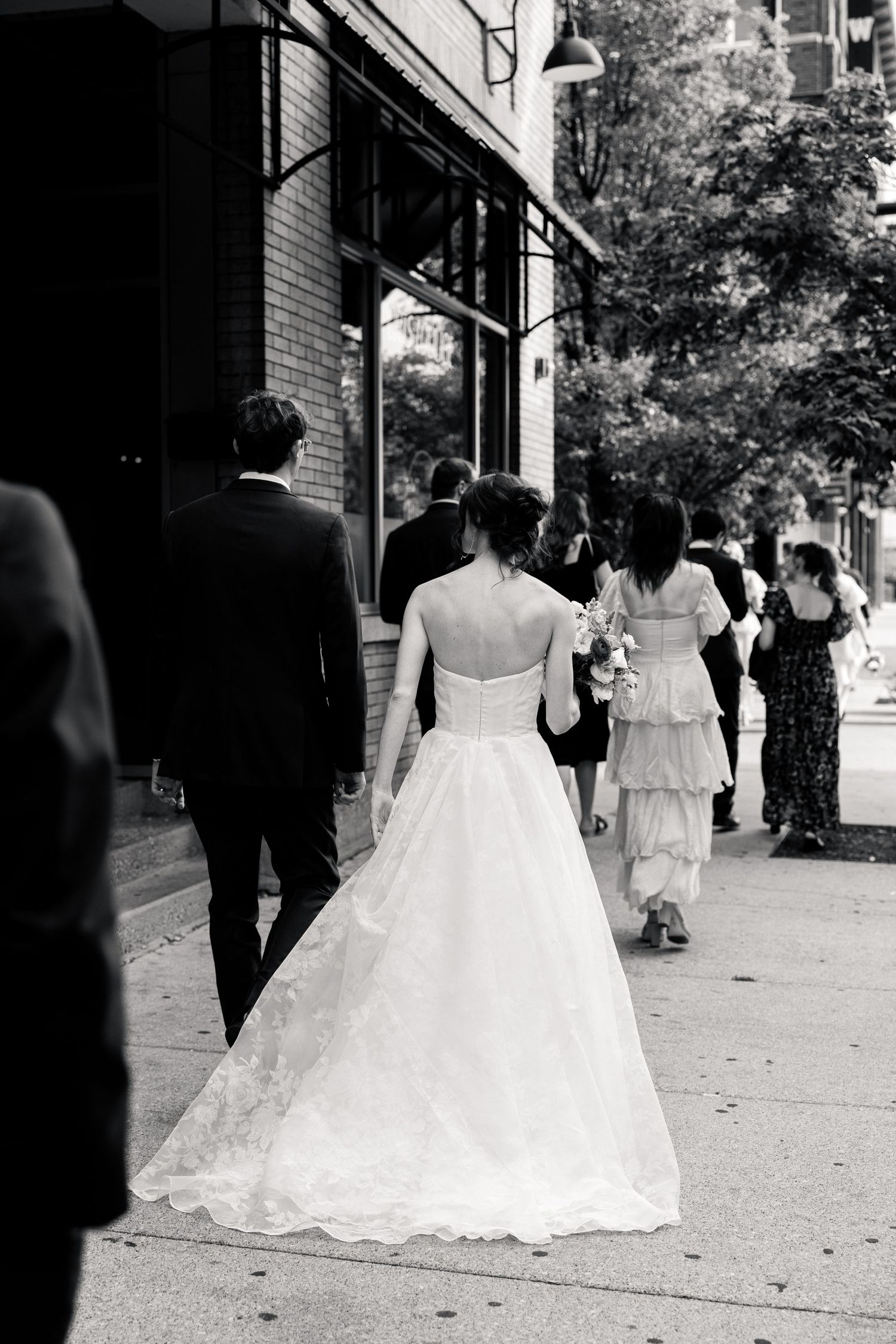 Black and white photo of bride in ball gown walking with groom and guests on city sidewalk.