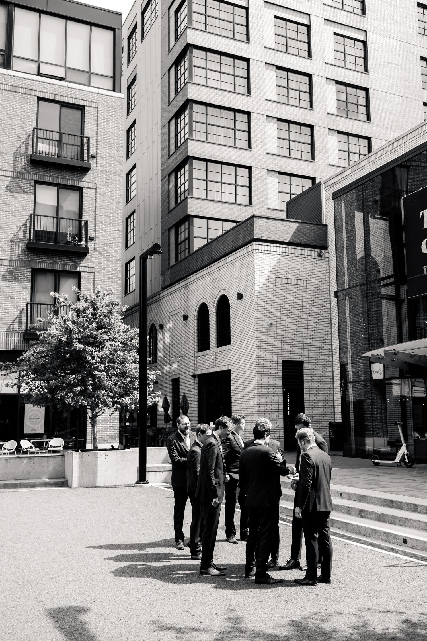 Black and white photo of groomsmen gathered in a circle outside modern brick buildings on a wedding day.