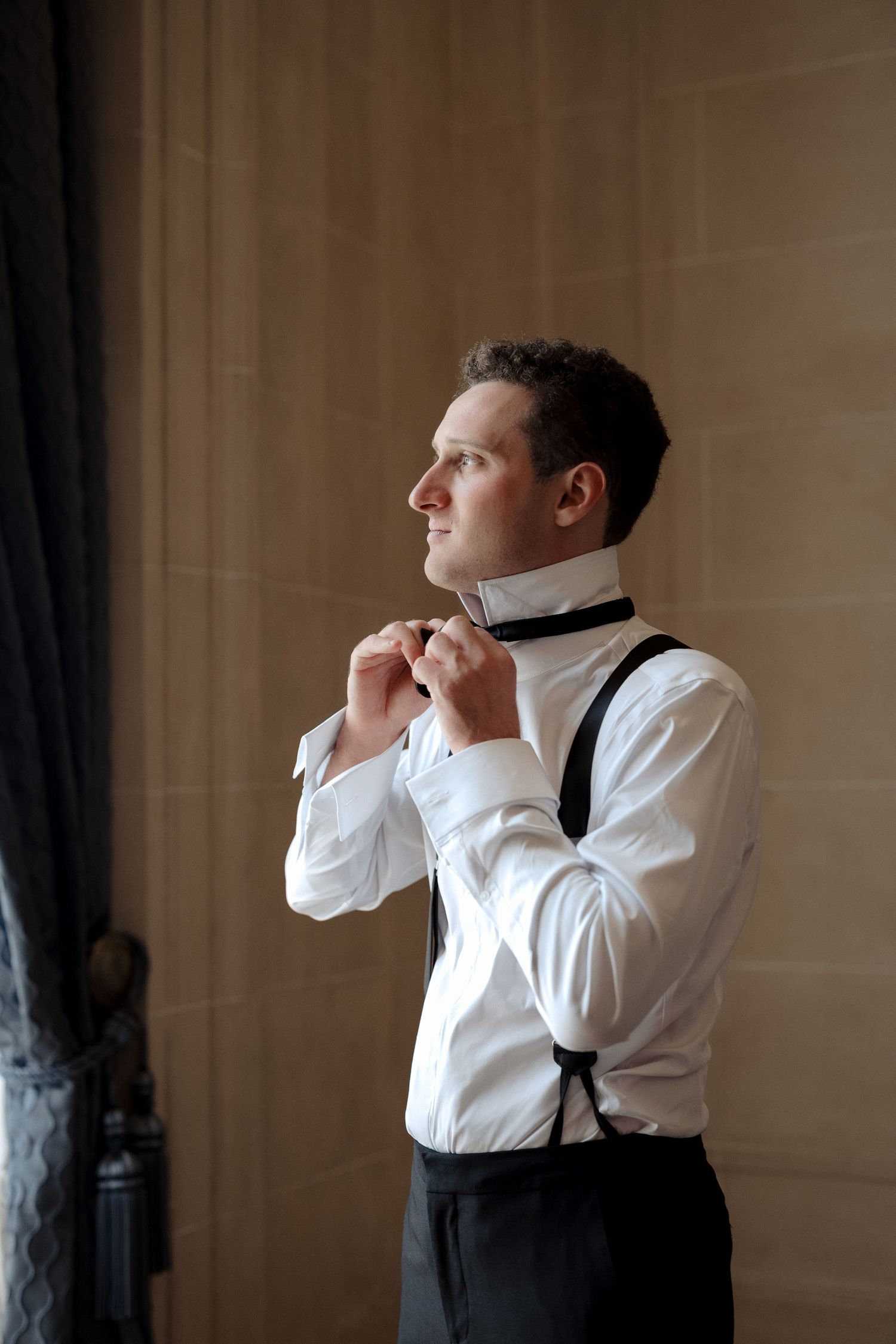 Groom in white dress shirt with black suspenders adjusts his bow tie while standing by a window.