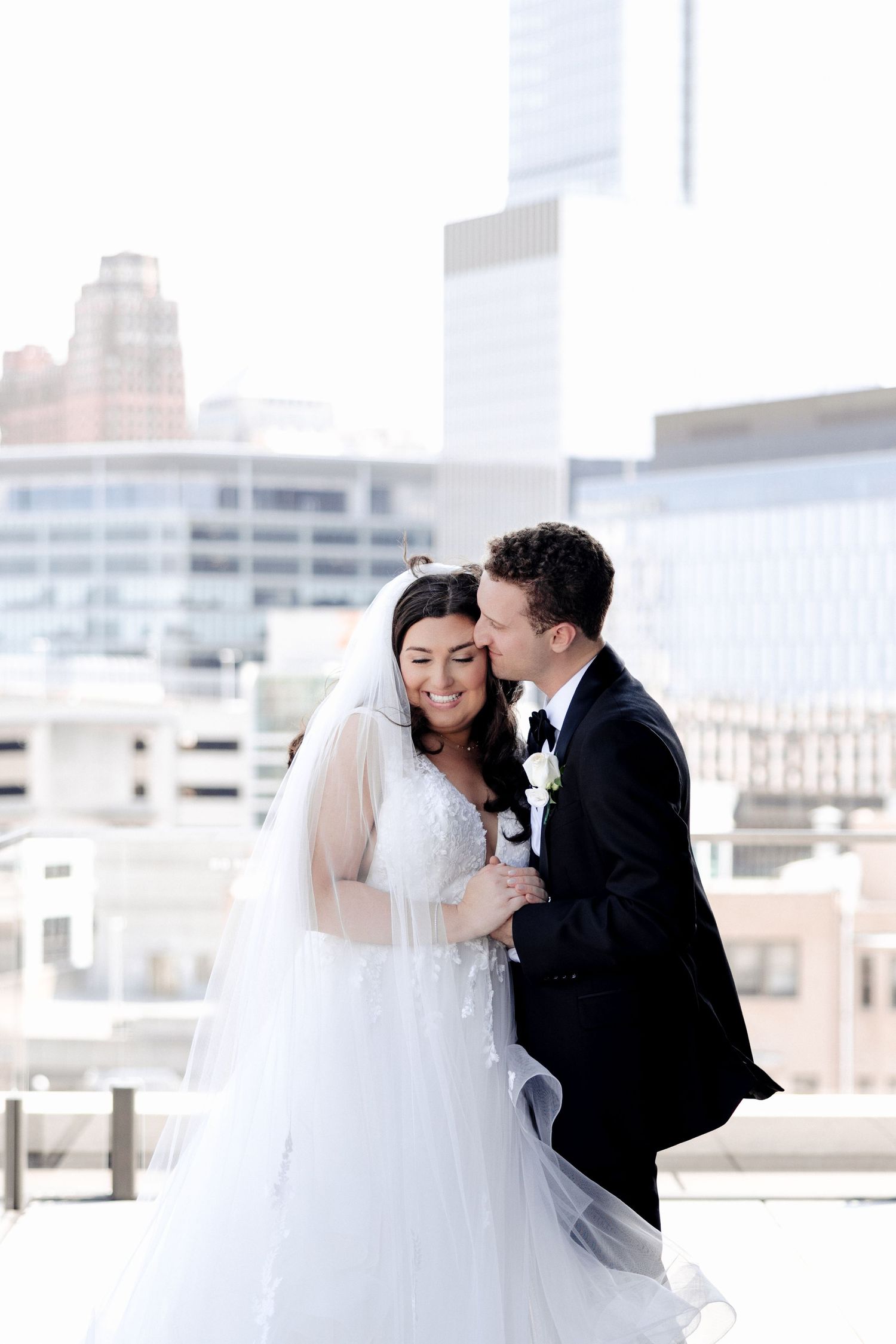 Groom kissing bride on cheek on rooftop with city skyline in background.