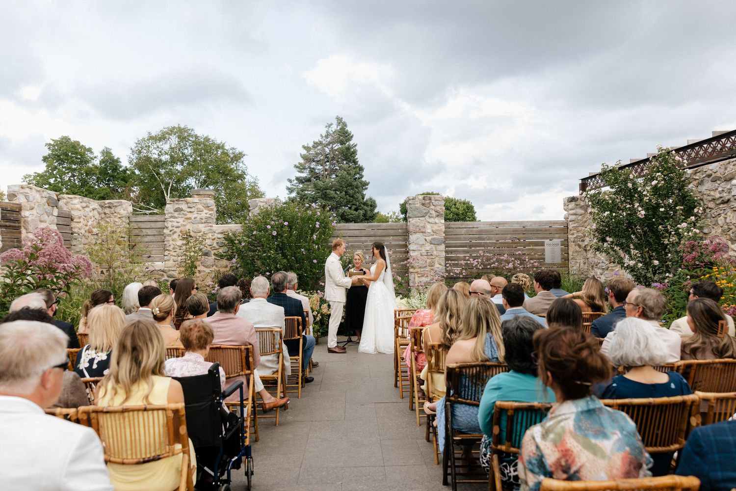 Outdoor wedding ceremony in a stone garden courtyard with seated guests watching couple exchange vows under cloudy sky.