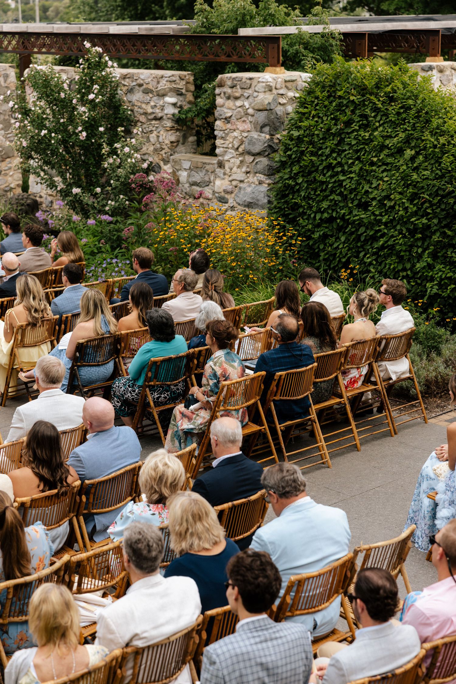 Guests seated in rattan chairs at an outdoor wedding ceremony surrounded by lush greenery and stone walls.