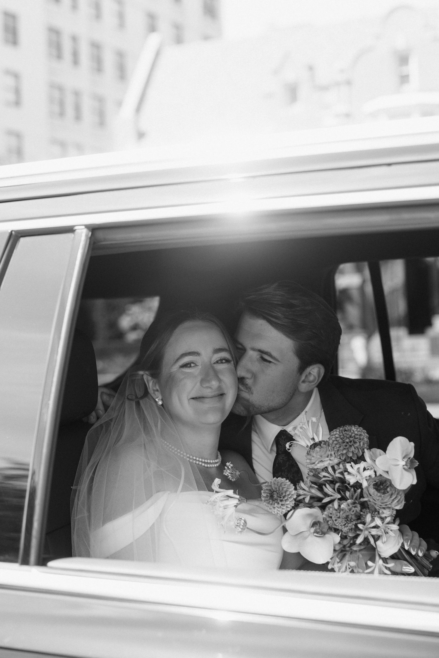 Black and white photo of a bride and groom sharing a kiss in a vintage car, bride holding a bouquet.