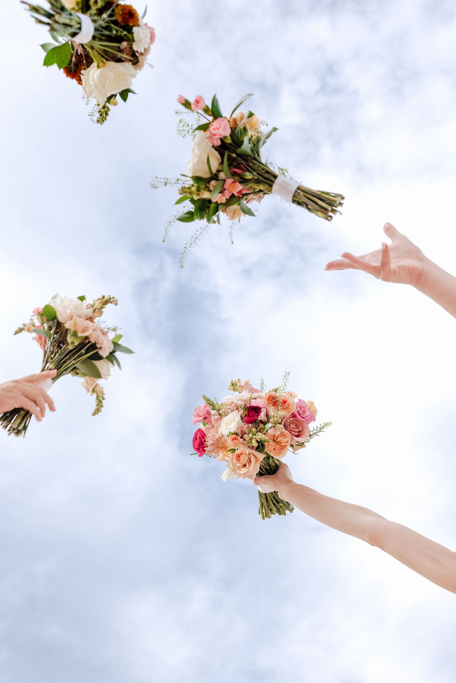 Bridesmaids toss colorful rose bouquets into cloudy sky in upward view wedding photo.