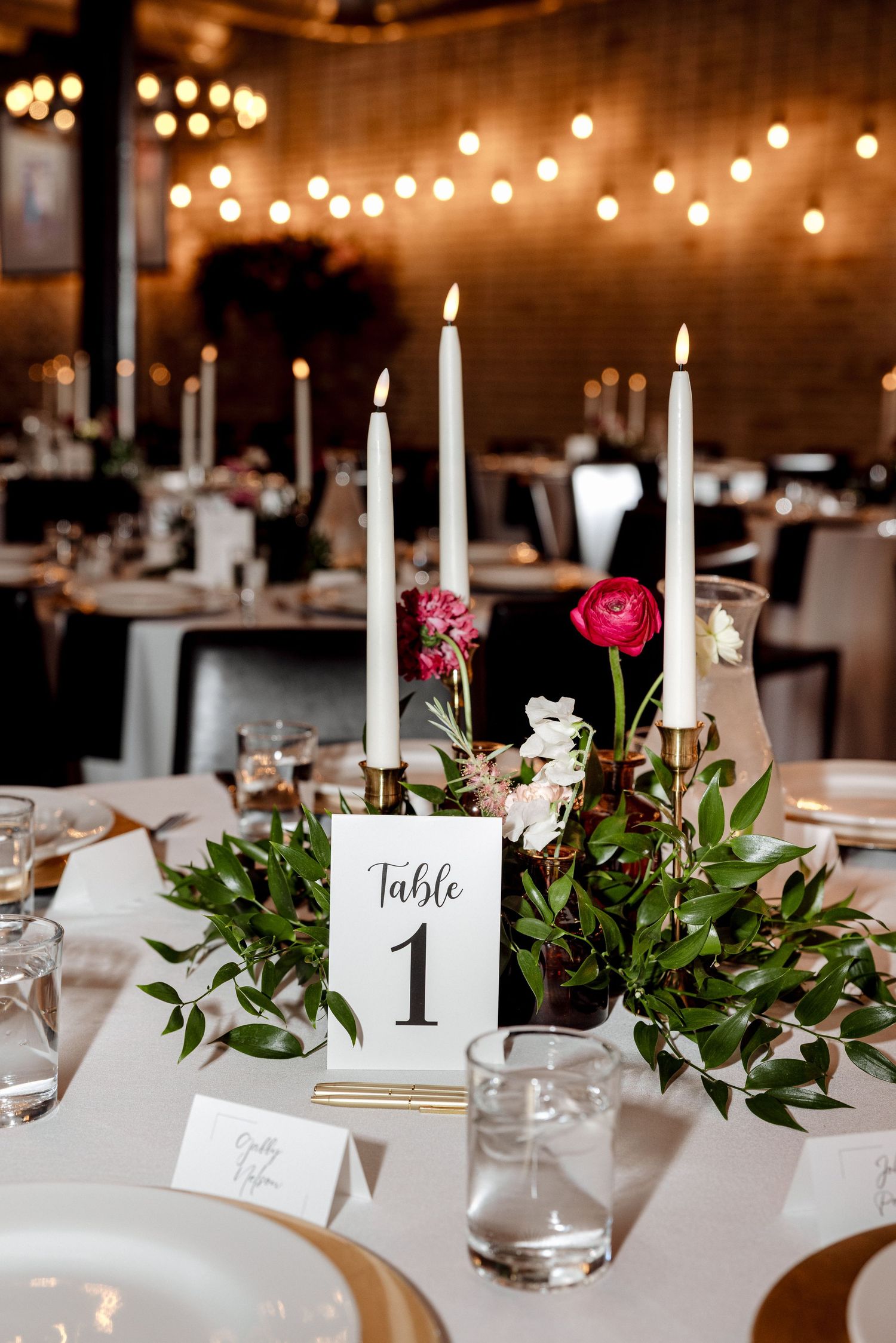 Wedding Table 1 centerpiece with white taper candles, pink roses, and greenery in warm reception hall.