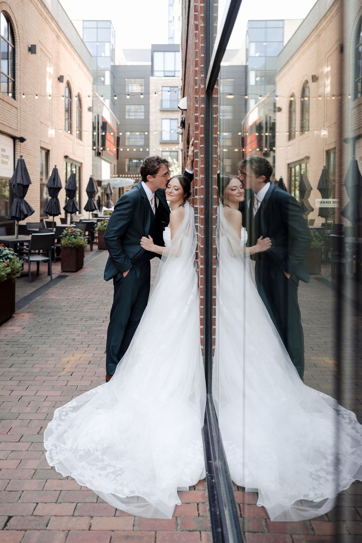 Bride in a long lace wedding gown and groom embrace near a reflective glass wall in an outdoor courtyard.
