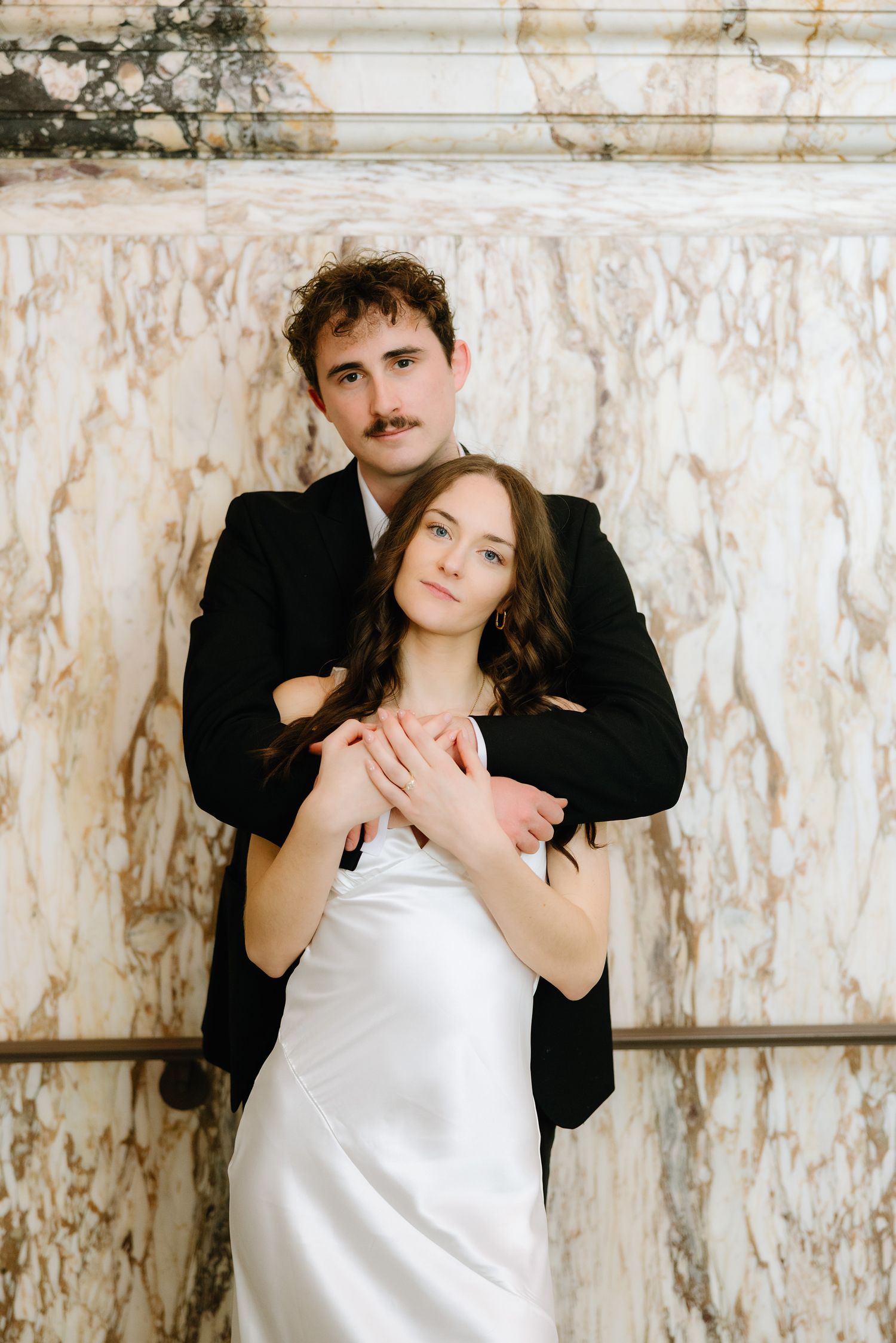Groom in black suit embracing bride in white satin dress against marble wall.