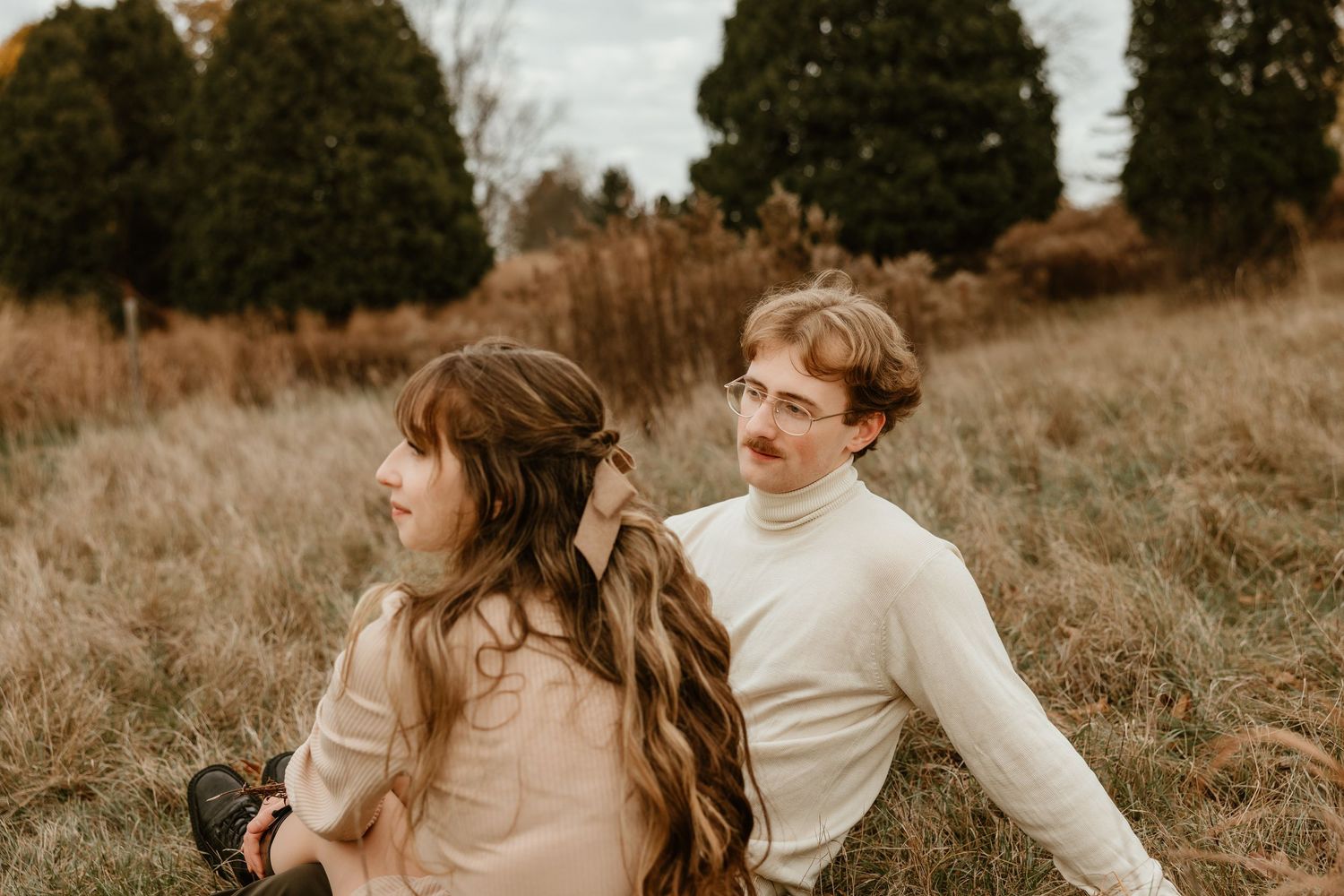 A couple sits in a grassy field, the woman looking away as the man gazes at her adoringly.