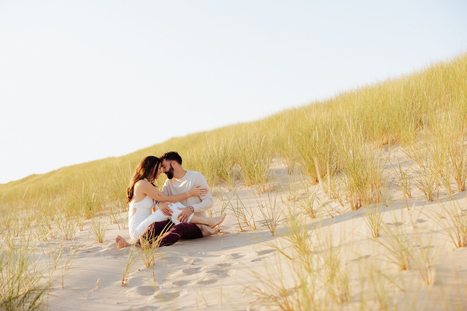 Couple sitting together on sandy beach dunes surrounded by tall grass under a clear sky.
