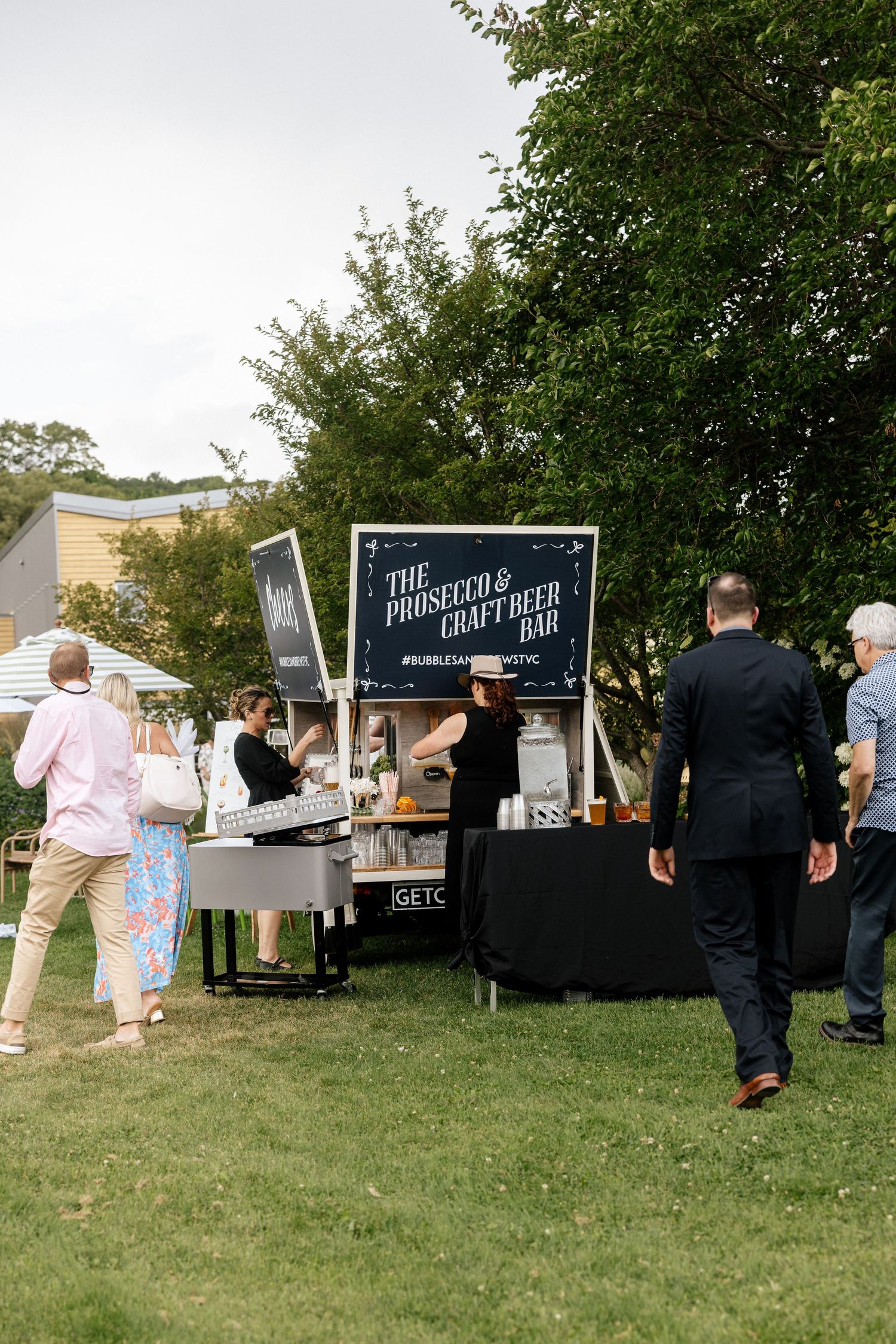 Outdoor prosecco and craft beer bar setup at a garden event with guests browsing on a sunny day.