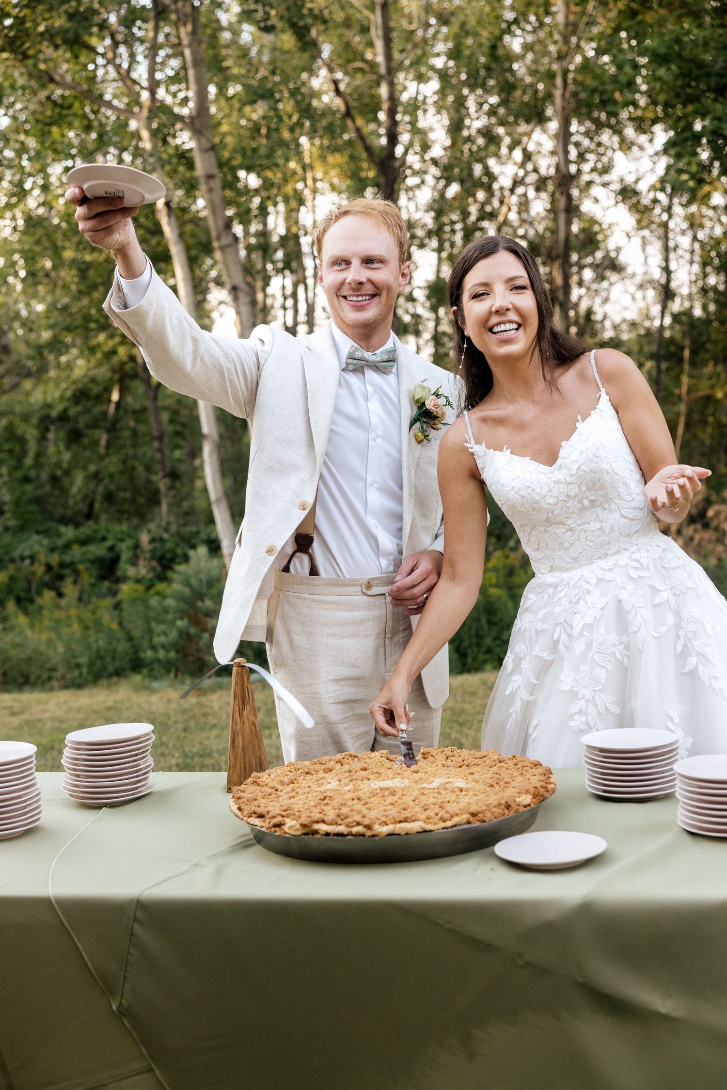 Smiling bride and groom cutting a rustic pie at their outdoor wedding reception surrounded by trees.