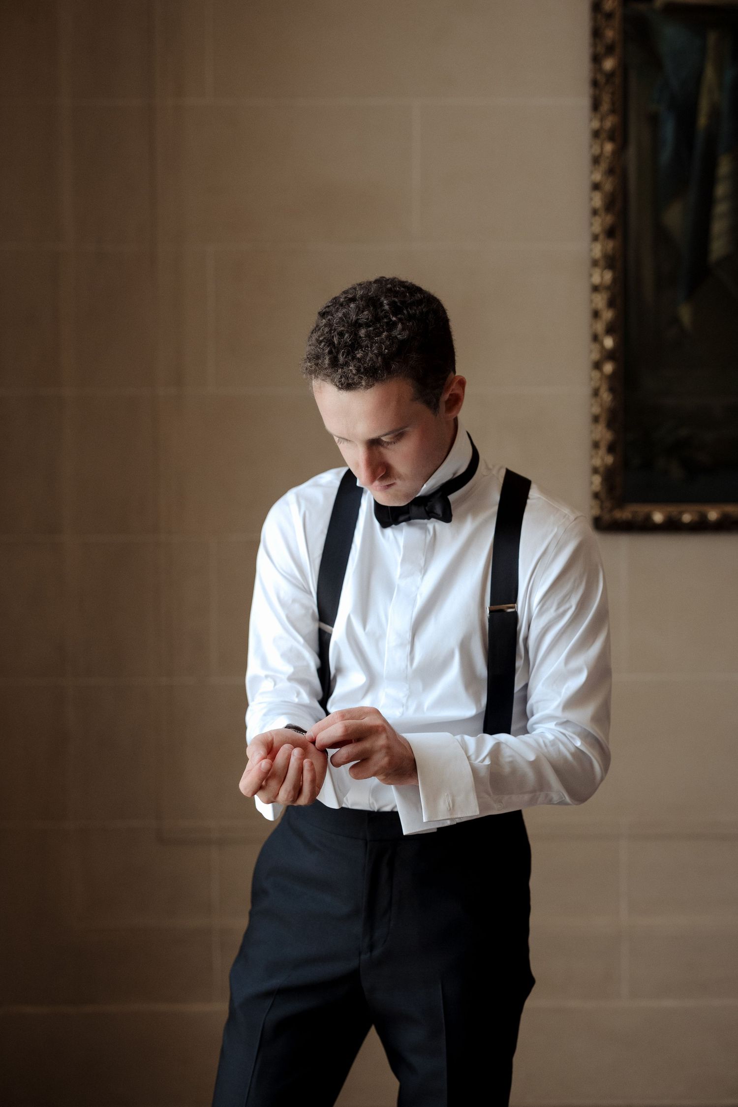 Groom in white dress shirt with black suspenders and bow tie fastens his cufflinks before wedding ceremony.
