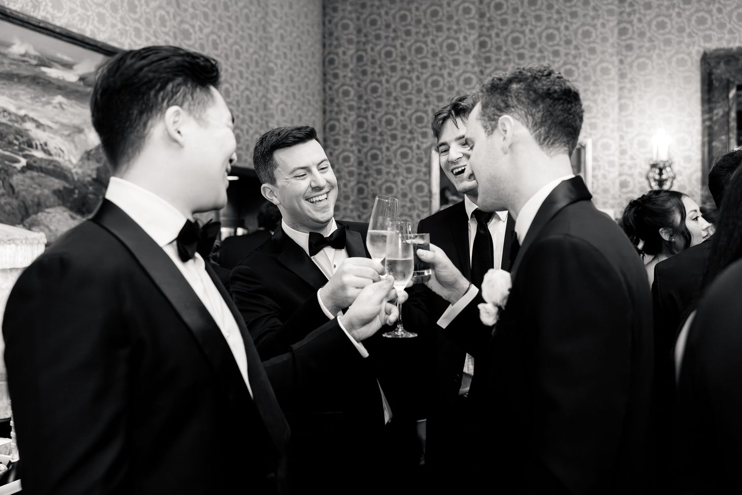 Four men in black tuxedos laughing and toasting champagne at a wedding celebration.