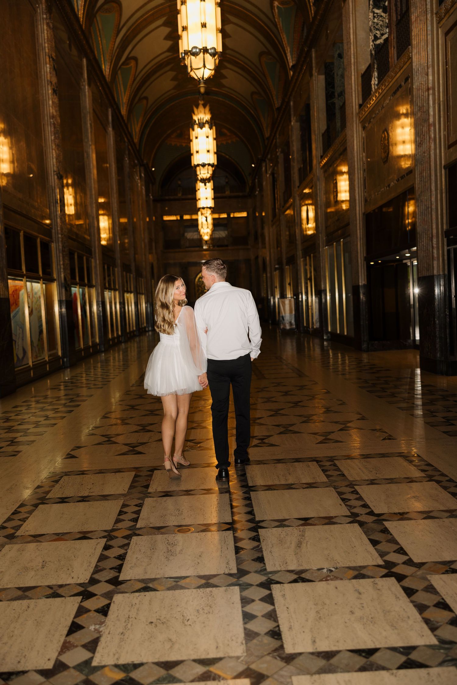 Couple holding hands walking through an ornate golden hallway with chandeliers and marble floors.